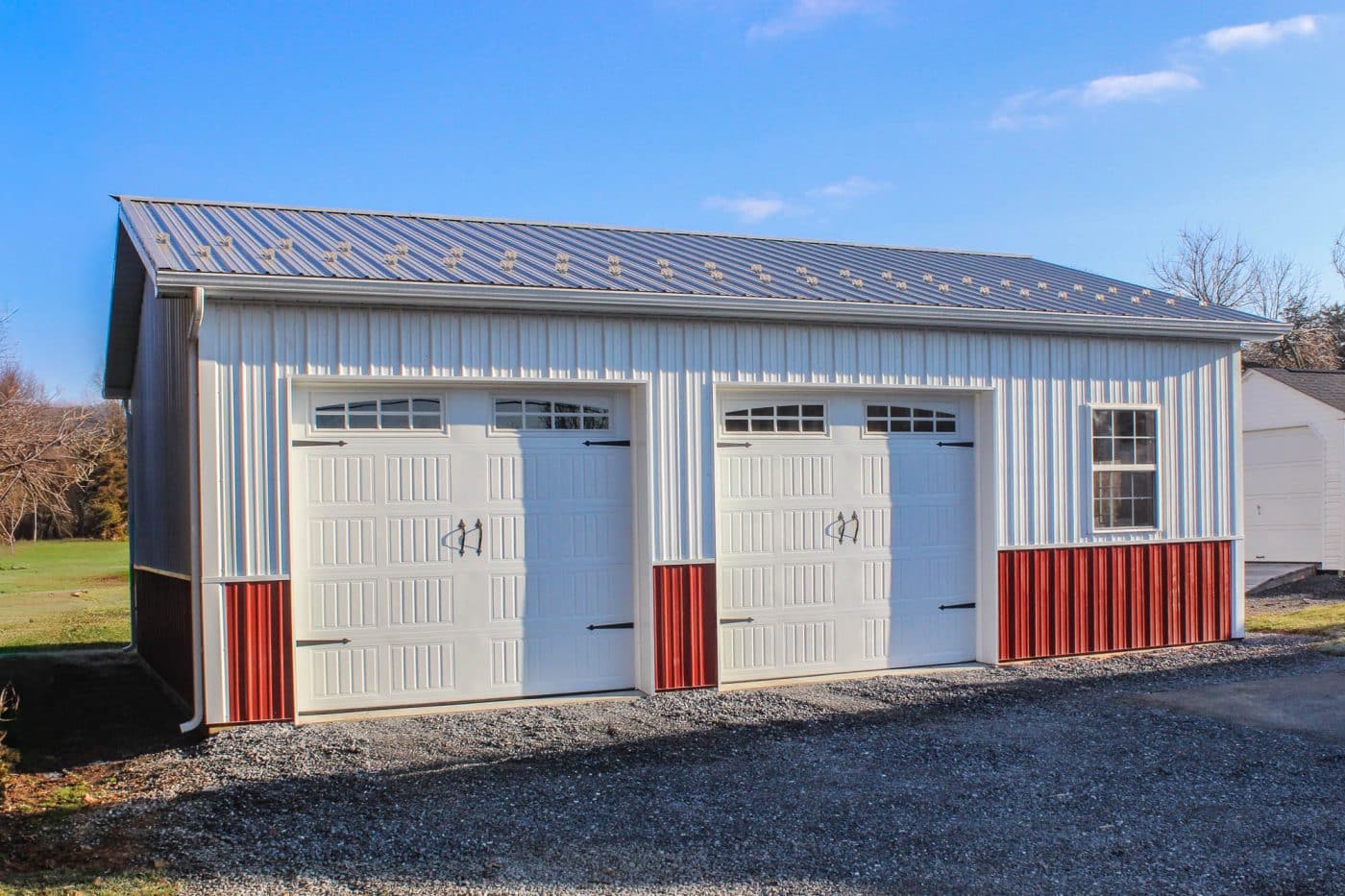 white and red post-frame garage with two overhead doors, metal roof, gravel driveway, and large side window.