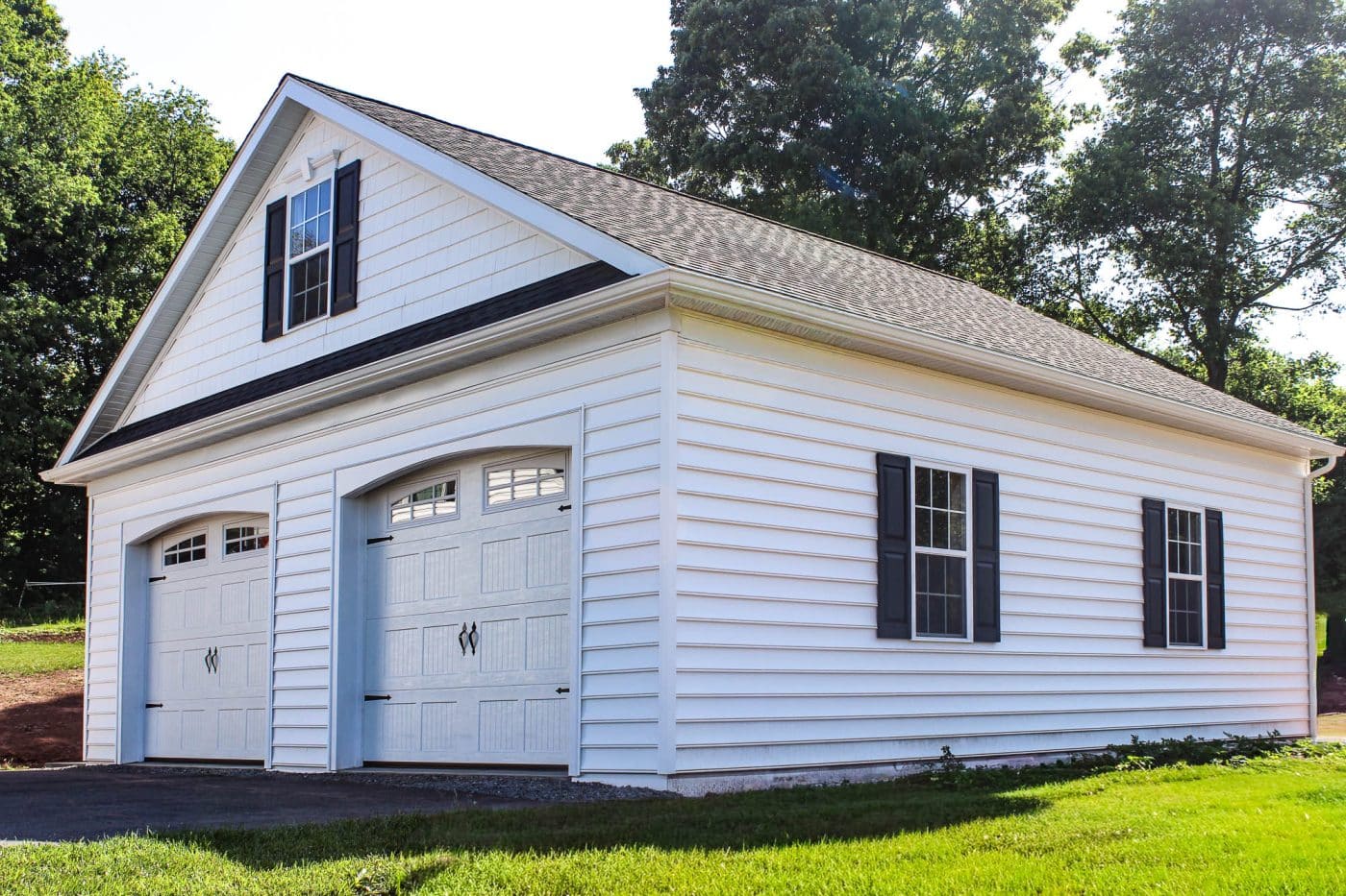 white two-bay pole garage with overhead doors, black shutters, upper window, and shingle roof beside a lawn.