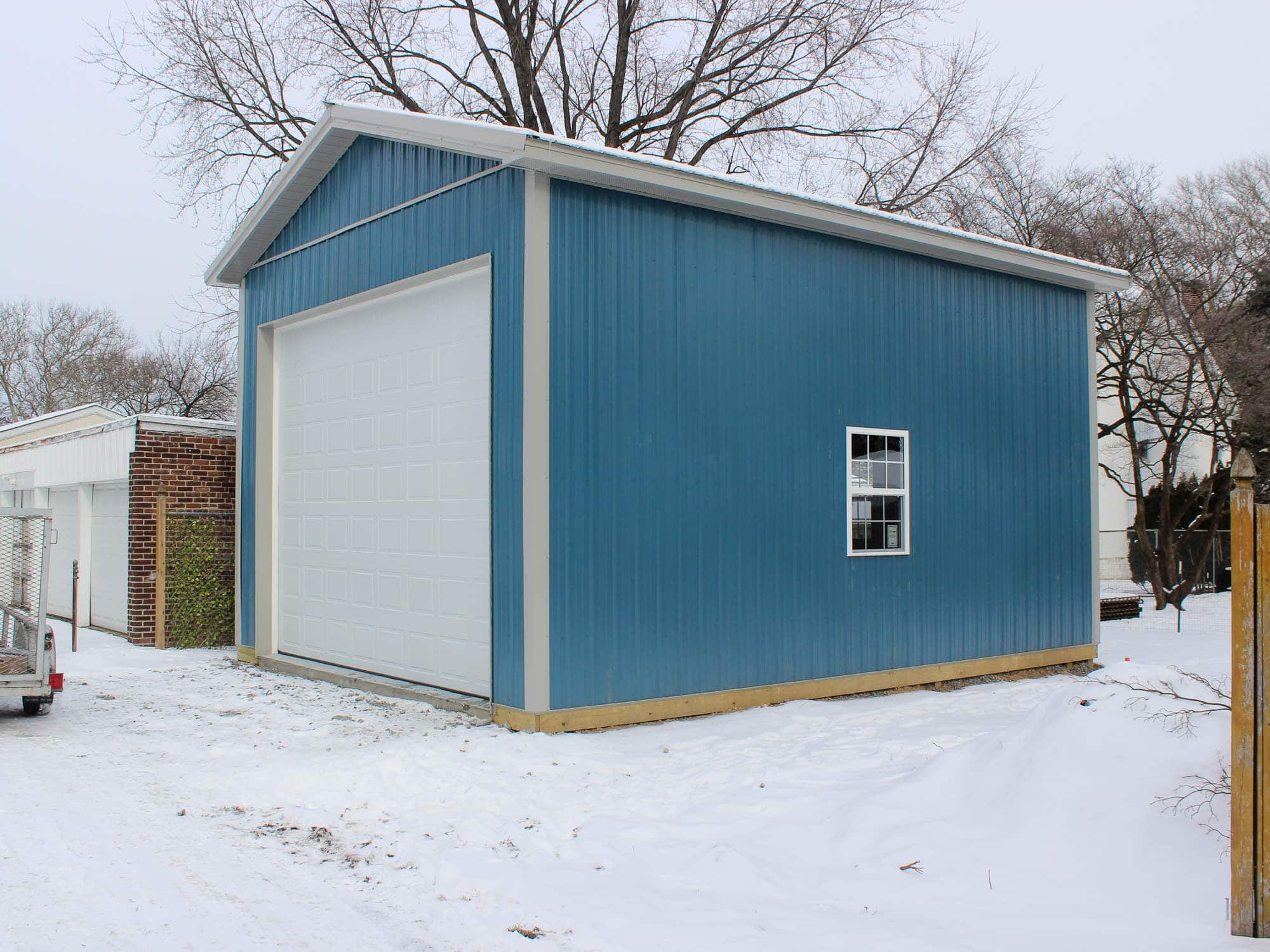 A blue metal garage with a white roll-up door and a side window. The building has a gable roof with white trim and is positioned on a snow-covered lot with trees and neighboring houses in the background.