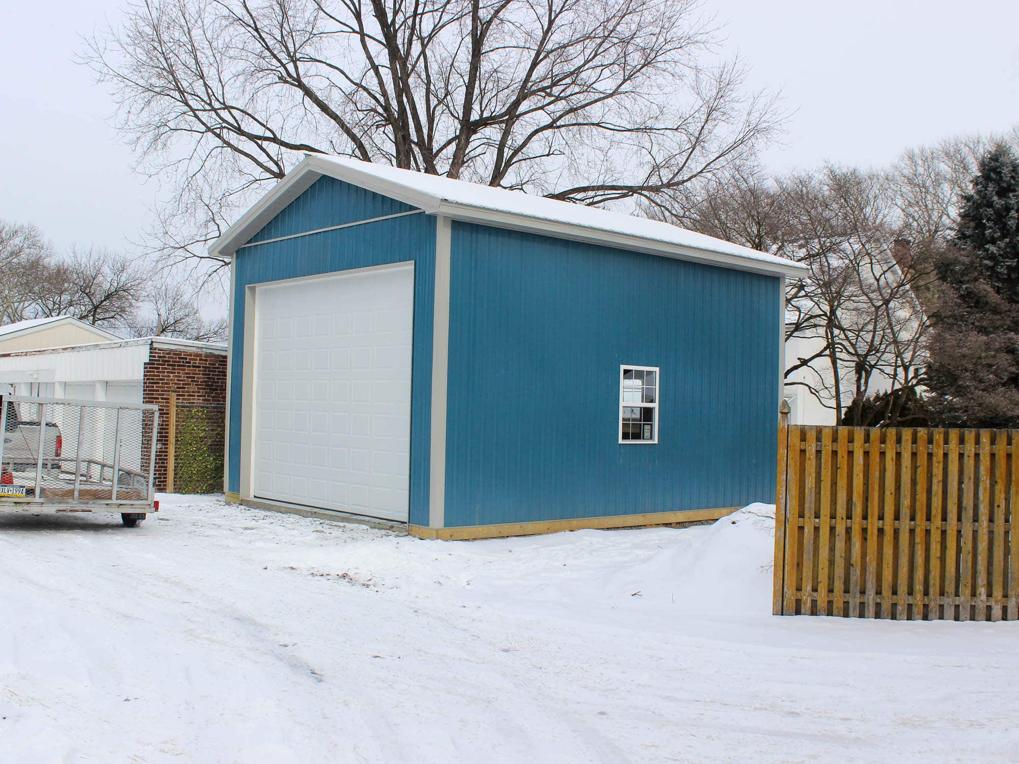 A blue metal garage with a large white roll-up door and a gable roof. The building is situated on a snow-covered lot with a wooden fence and neighboring houses visible in the background.