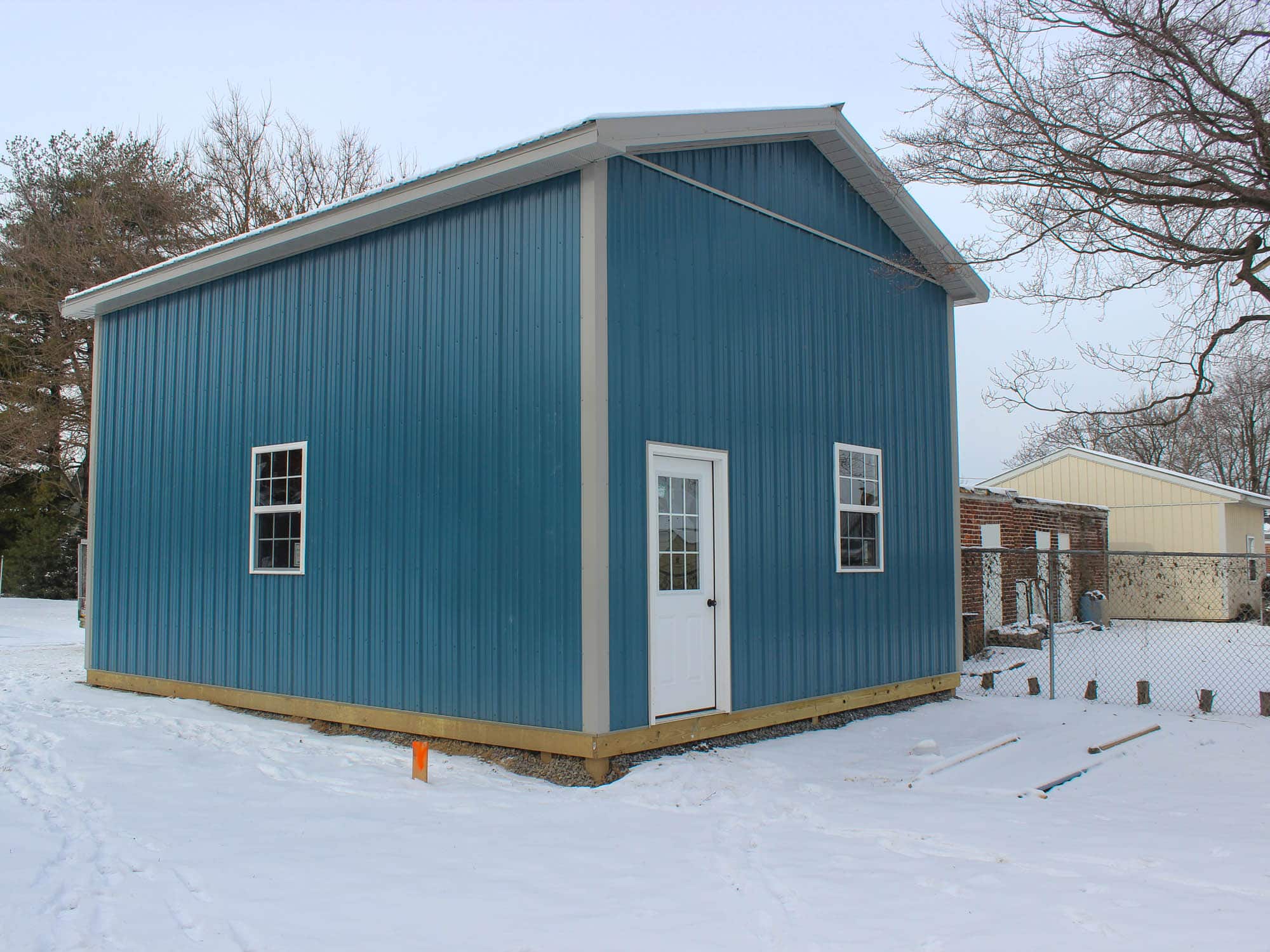 A blue metal building with a white side door and a window, positioned on a snow-covered lot. The gable roof features white trim, and the structure is surrounded by residential houses and trees.