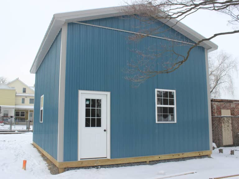 a blue metal garage with a white side door and a small window, set on a snowy lot with residential homes and trees in the background. the building has a gable roof with white trim and sits on a raised concrete foundation.