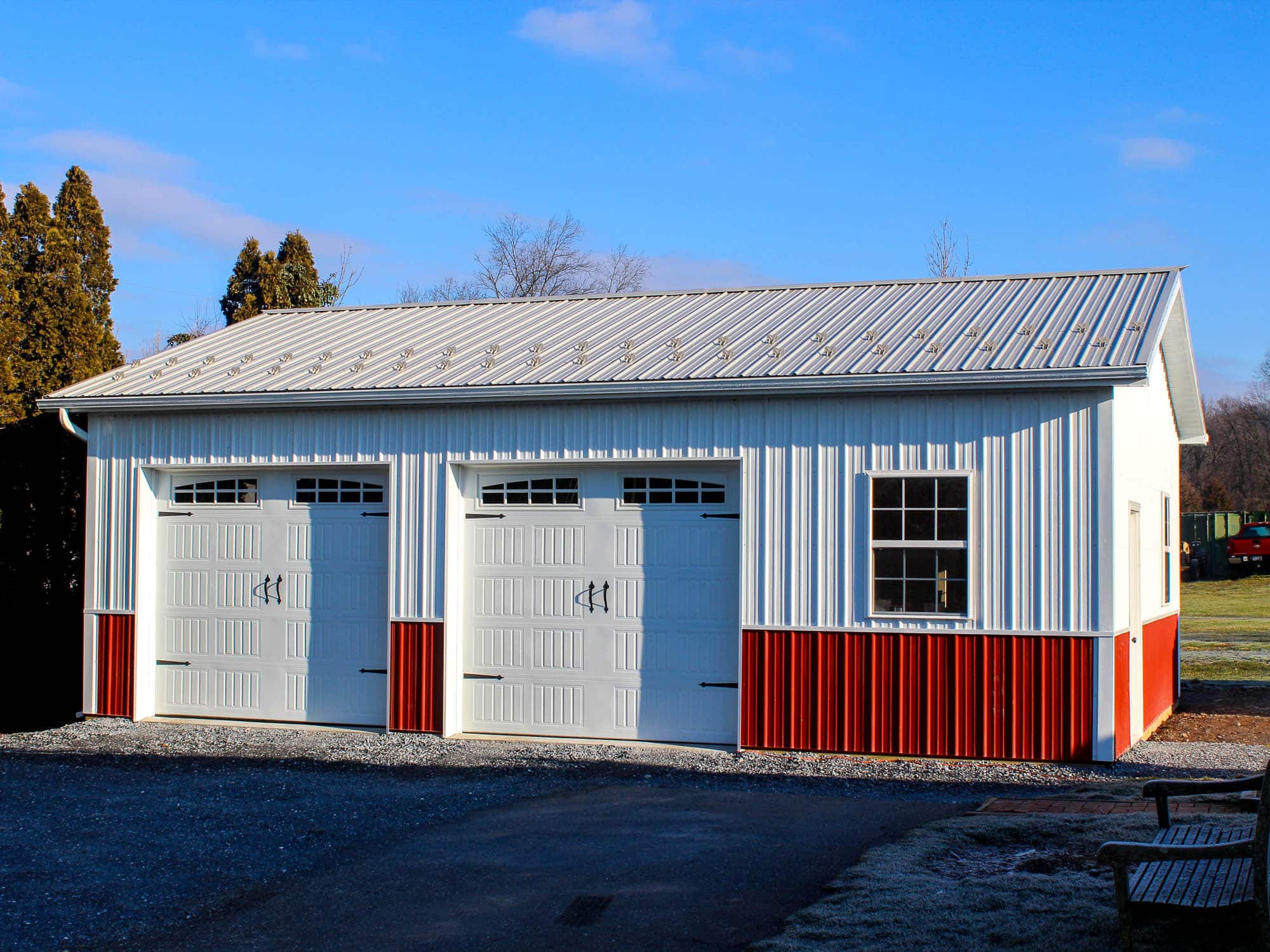A white metal garage with red wainscoting, two white roll-up garage doors with small windows, and a gray metal roof. The structure sits on a gravel driveway with grass and trees in the background.