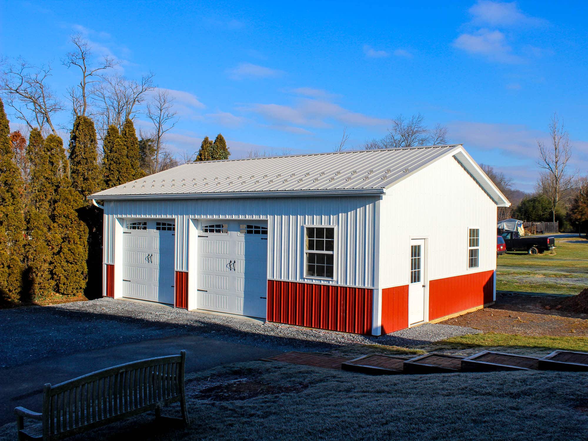 An angled view of a white and red metal garage with two roll-up doors and a gray metal roof. The structure is surrounded by gravel and a well-maintained lawn with trees in the background.