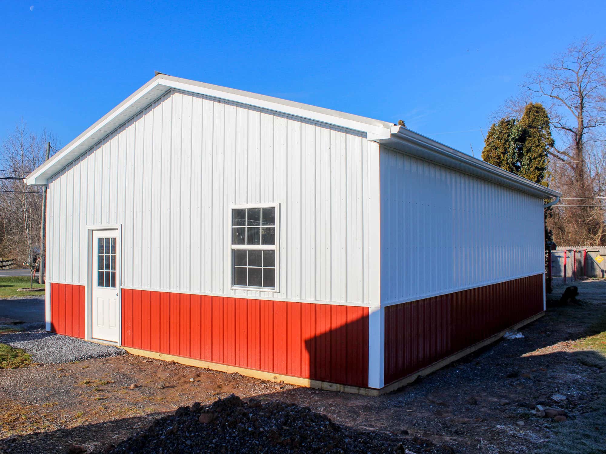 A side view of a white and red metal garage with a gray metal roof. The structure features a small window and sits on a gravel lot surrounded by trees and grass.
