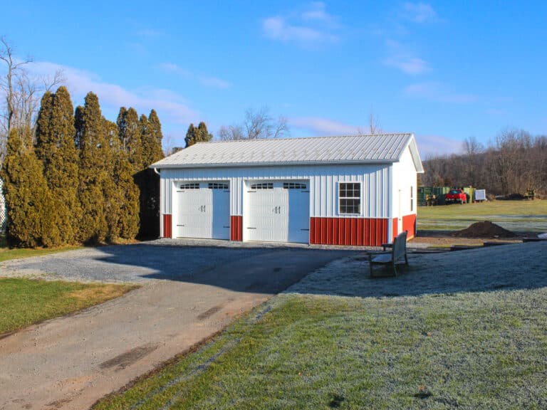 a white metal garage with red wainscoting, two white roll-up garage doors with small windows, and a gray metal roof. the structure sits on a gravel driveway with grass and trees in the background.