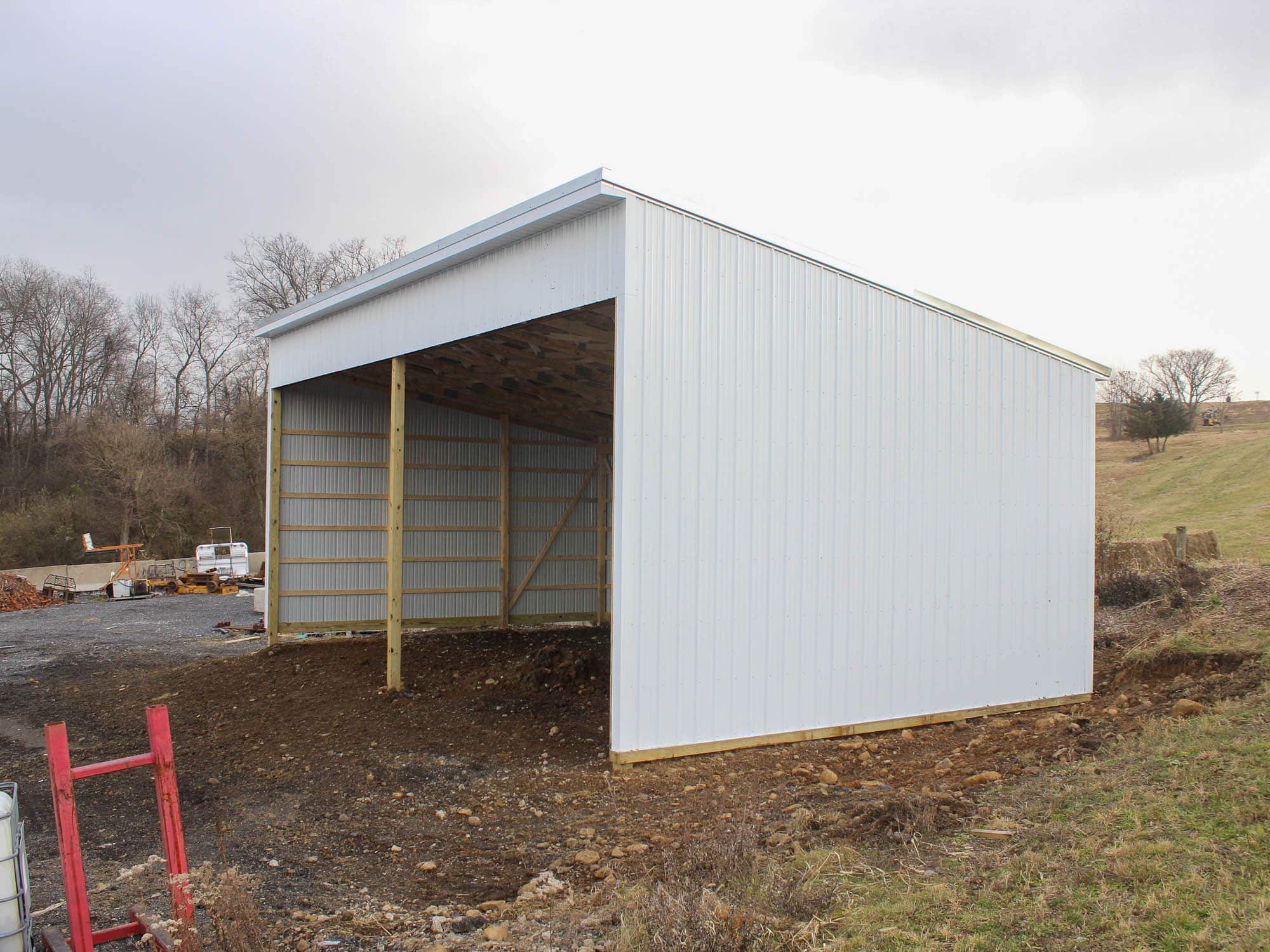 A white open-faced metal storage shed with a sloped roof. The shed is positioned on a dirt lot with a red construction tool visible nearby.