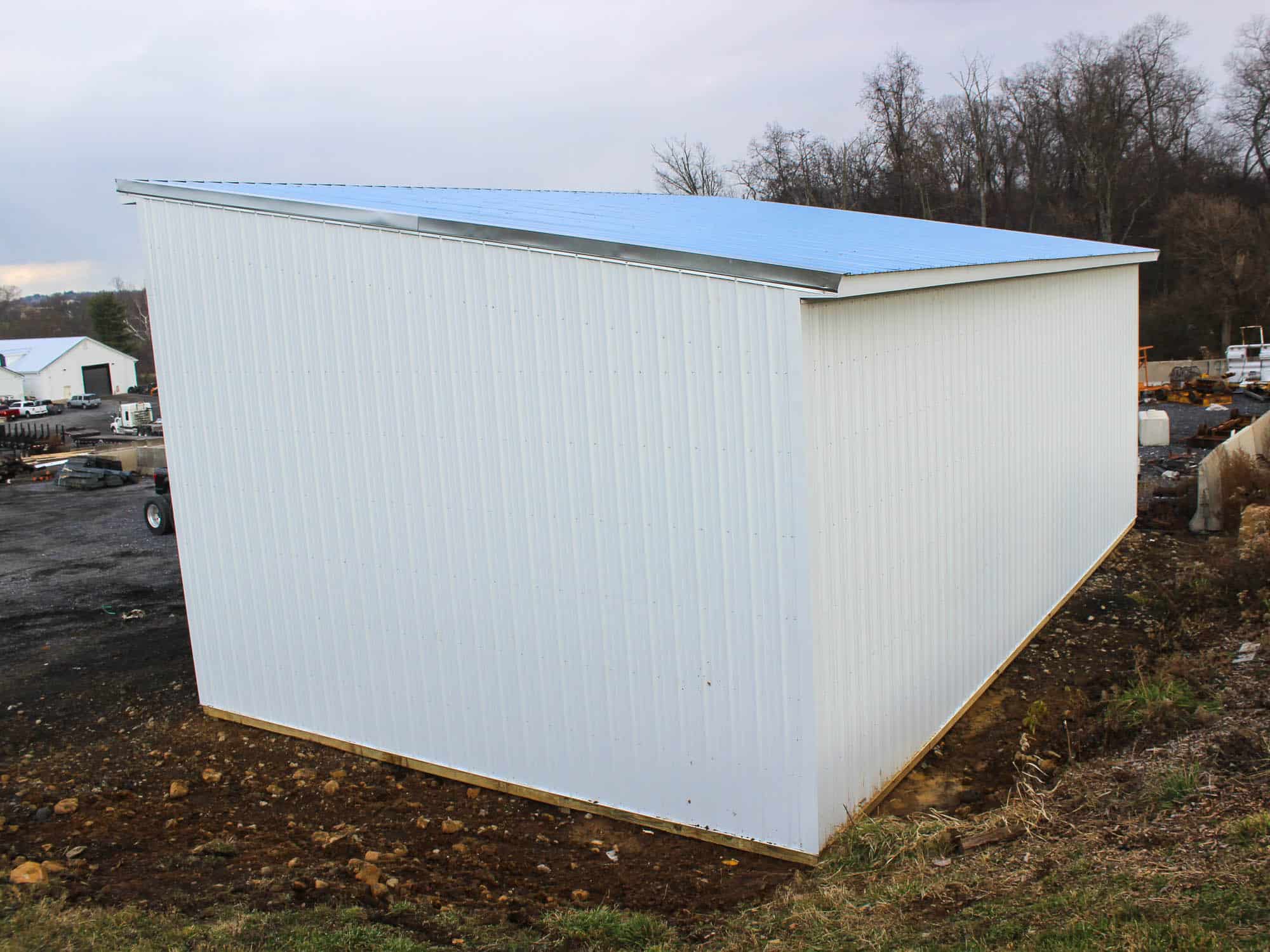 A small white lean-to shed with a gray metal roof. The shed is positioned on a dirt lot with trees and a clear sky in the background.