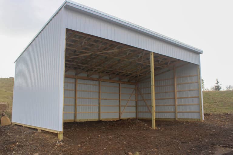 a large open-sided metal pole barn with white siding, a gray metal roof, and exposed wooden framing. the barn is set on a gravel lot with an open field and a clear sky in the background.