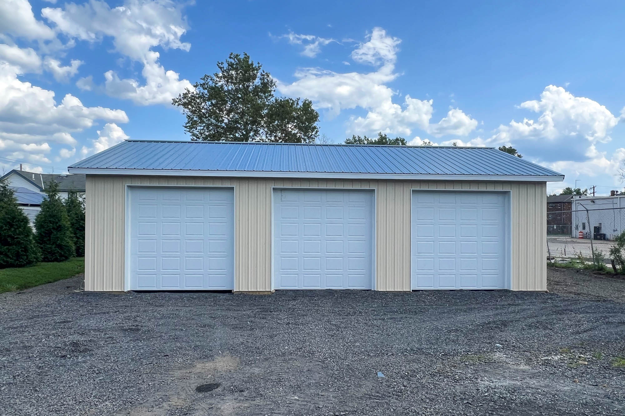 A side view of a beige metal garage with a gray gable roof. The structure sits on a gravel lot with dirt and grass surrounding the base.
