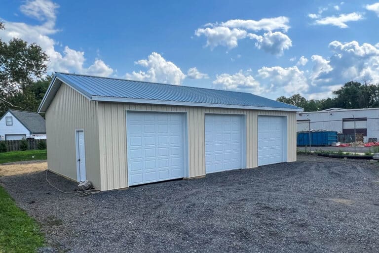 a front view of a beige metal garage with a gray metal roof and two white roll-up doors. the garage is situated on a gravel lot under a blue sky with scattered clouds.