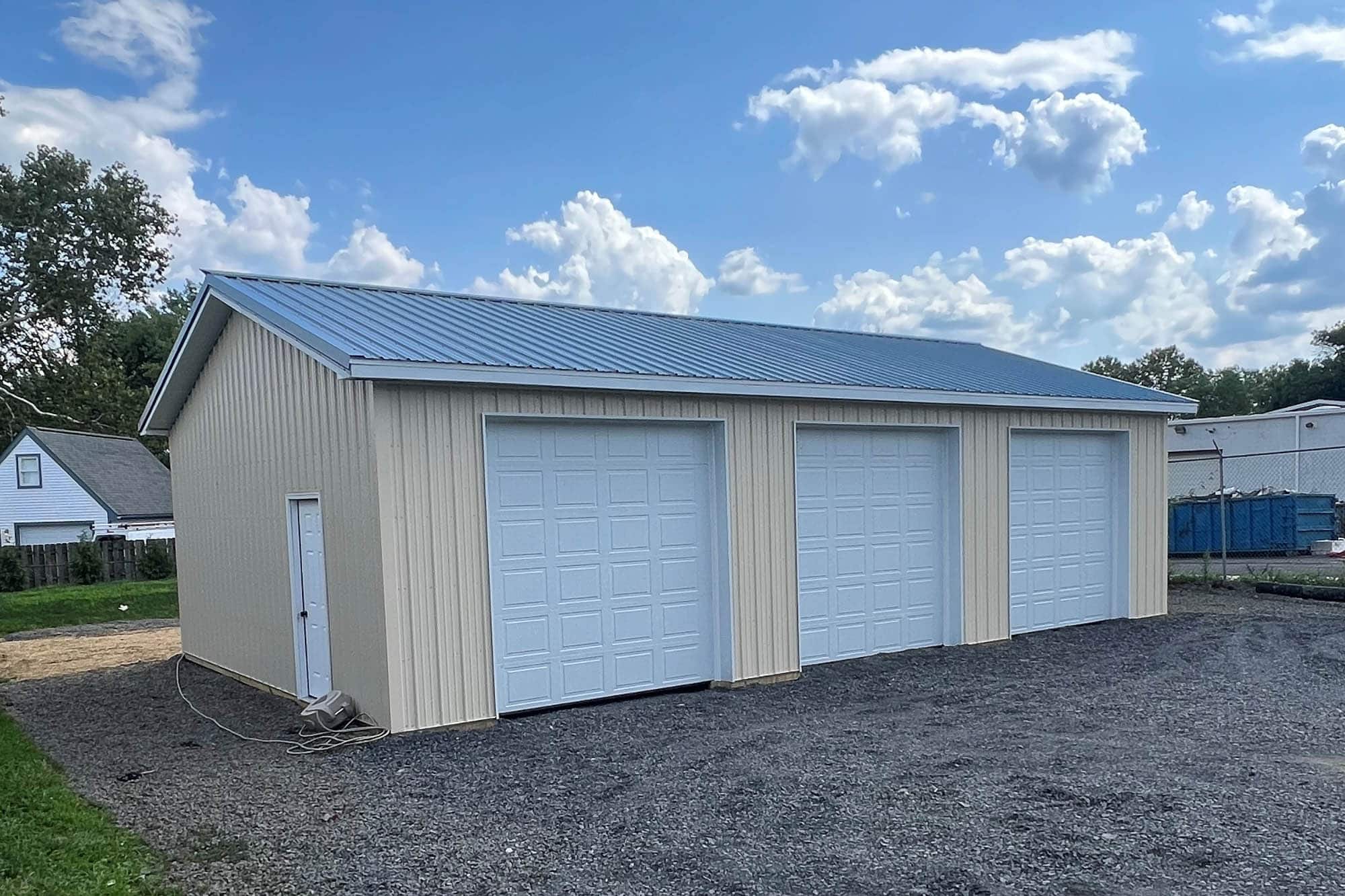 A beige metal garage with a gray metal roof and two white roll-up doors. The building sits on a gravel foundation with an open field and trees in the background.