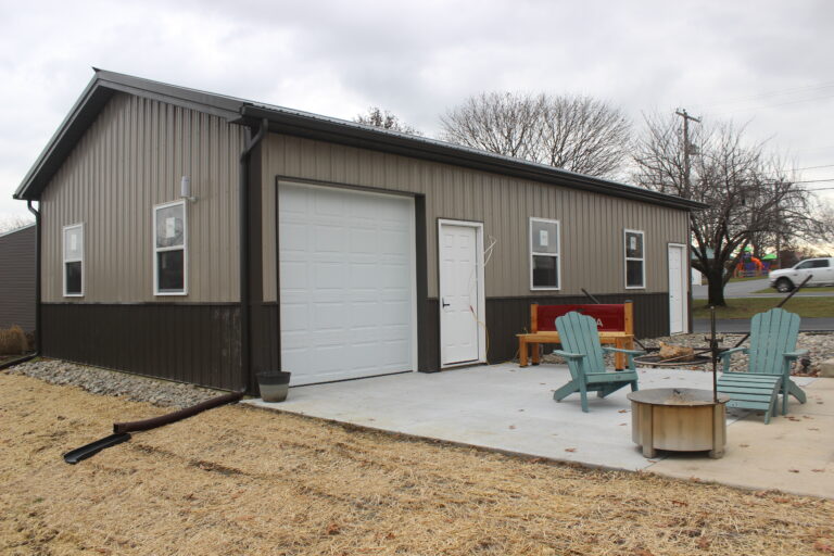 a gray metal garage with dark brown wainscoting, a white roll-up door, and an open side area with construction equipment nearby. the building sits on a concrete slab with a gravel pathway leading to the entrance.