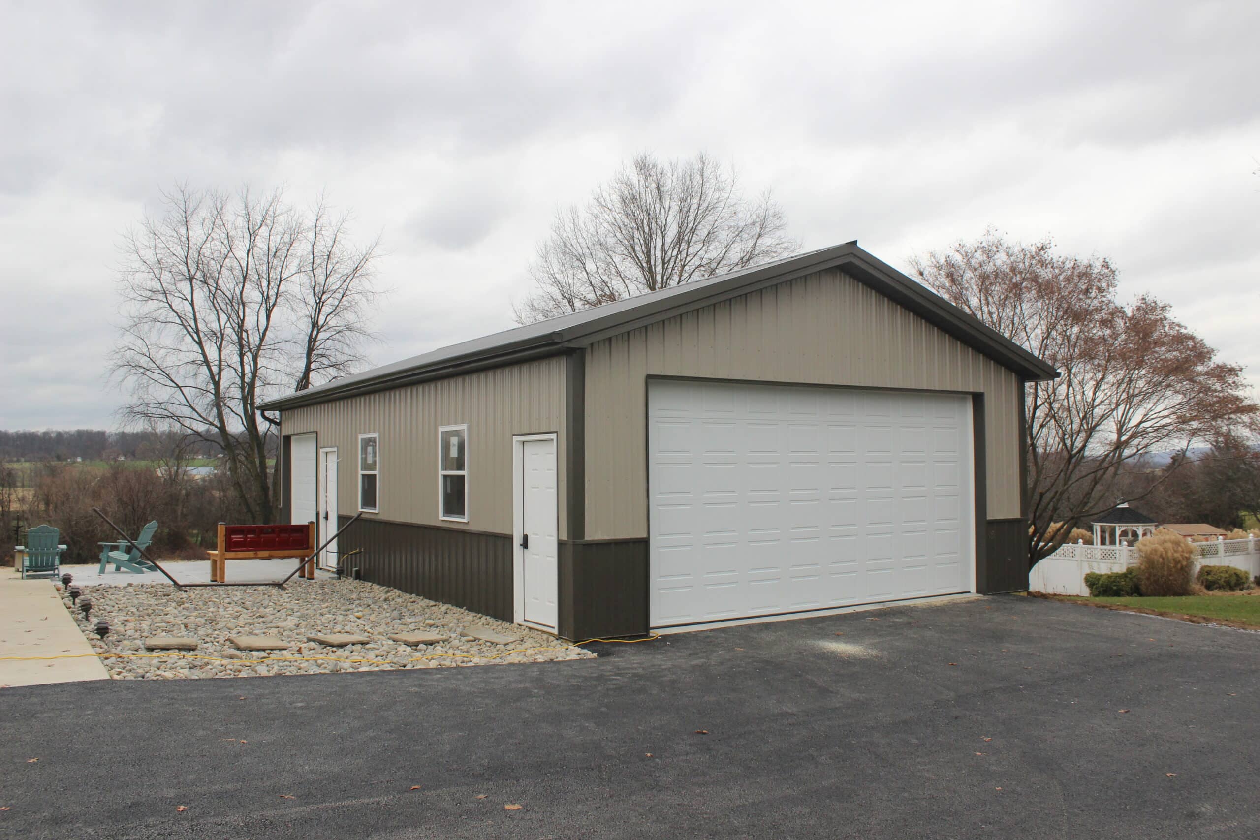 A side view of a gray metal garage with dark brown wainscoting, a white roll-up door, and a gable roof. The building sits on a concrete foundation with trees and a residential area nearby.