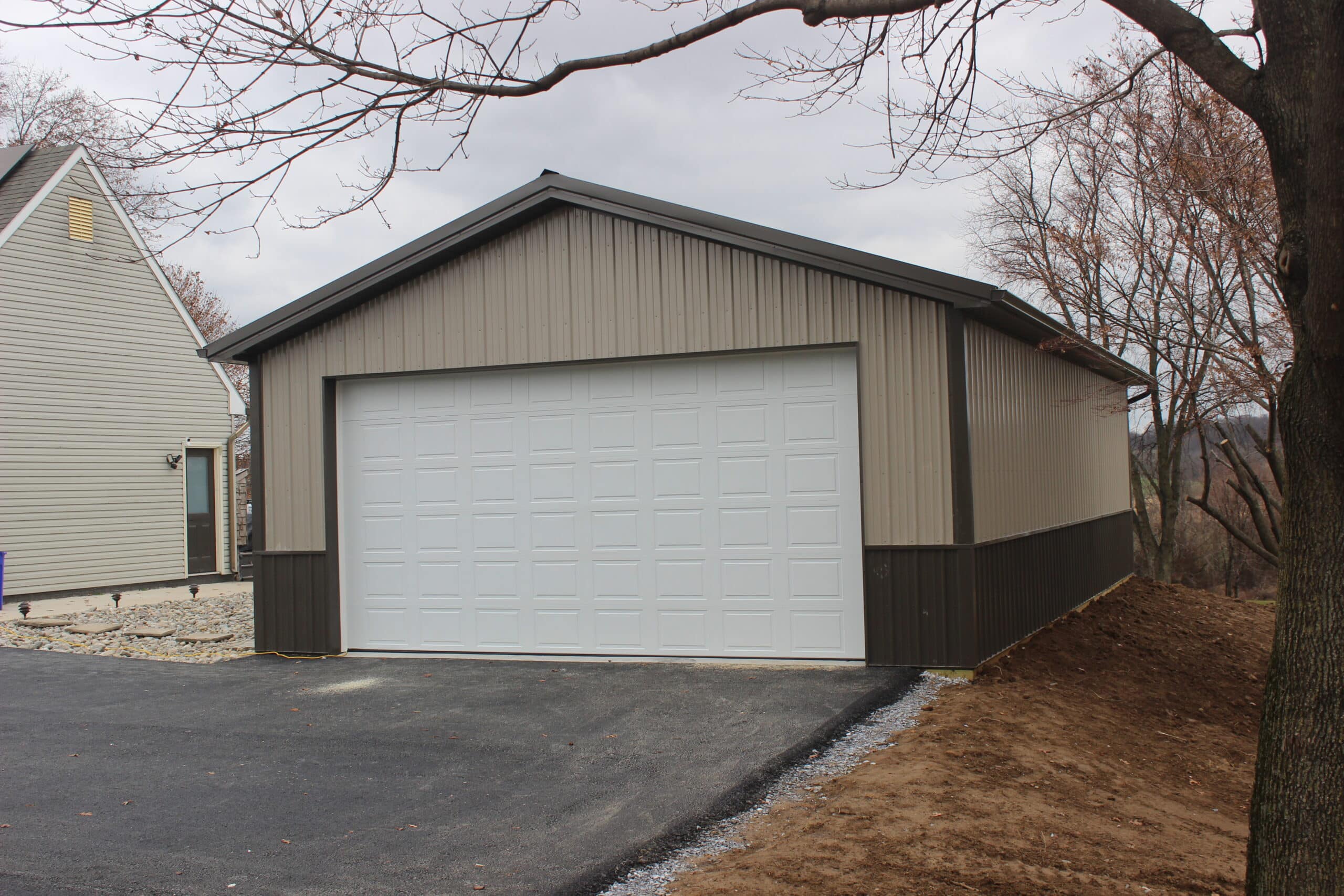 A gray metal garage with dark brown wainscoting, a large white roll-up garage door, and a gable roof with dark trim. The structure sits on a paved driveway with trees and a neighboring building in the background.