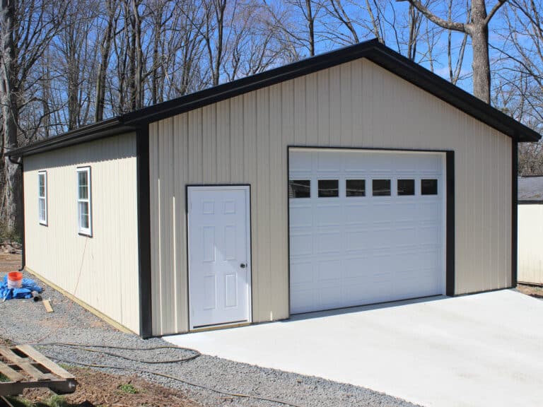 a beige metal garage with dark brown trim, a white roll-up garage door with small windows at the top, and a white side entry door. the building has a gable roof and is situated on a concrete driveway with trees in the background.