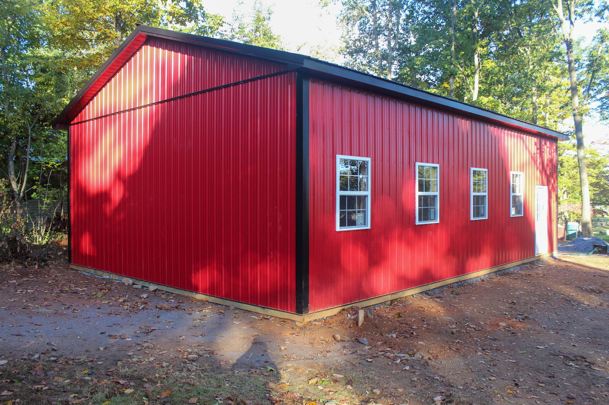 A red metal garage with a white roll-up door and a side entry door. The building features a gable roof and sits on a gravel lot surrounded by trees.
