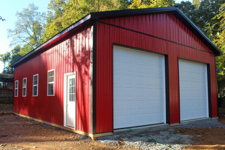 a red metal garage with a gable roof and a single white roll-up garage door. the structure is positioned on a gravel driveway with trees in the background.