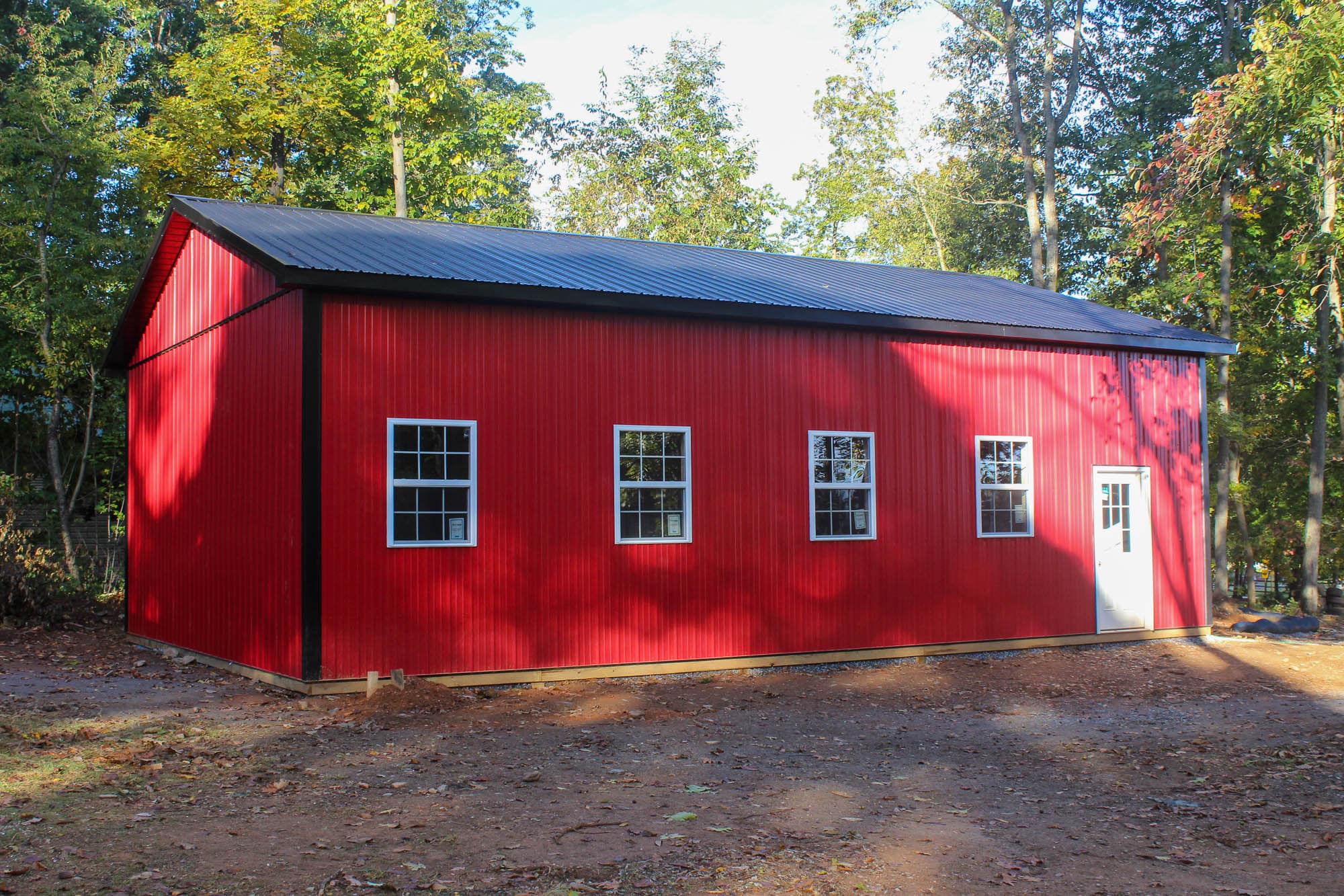 A red metal garage with a gray metal roof, featuring several white-framed windows and a side door. The building sits on a dirt lot surrounded by tall trees.
