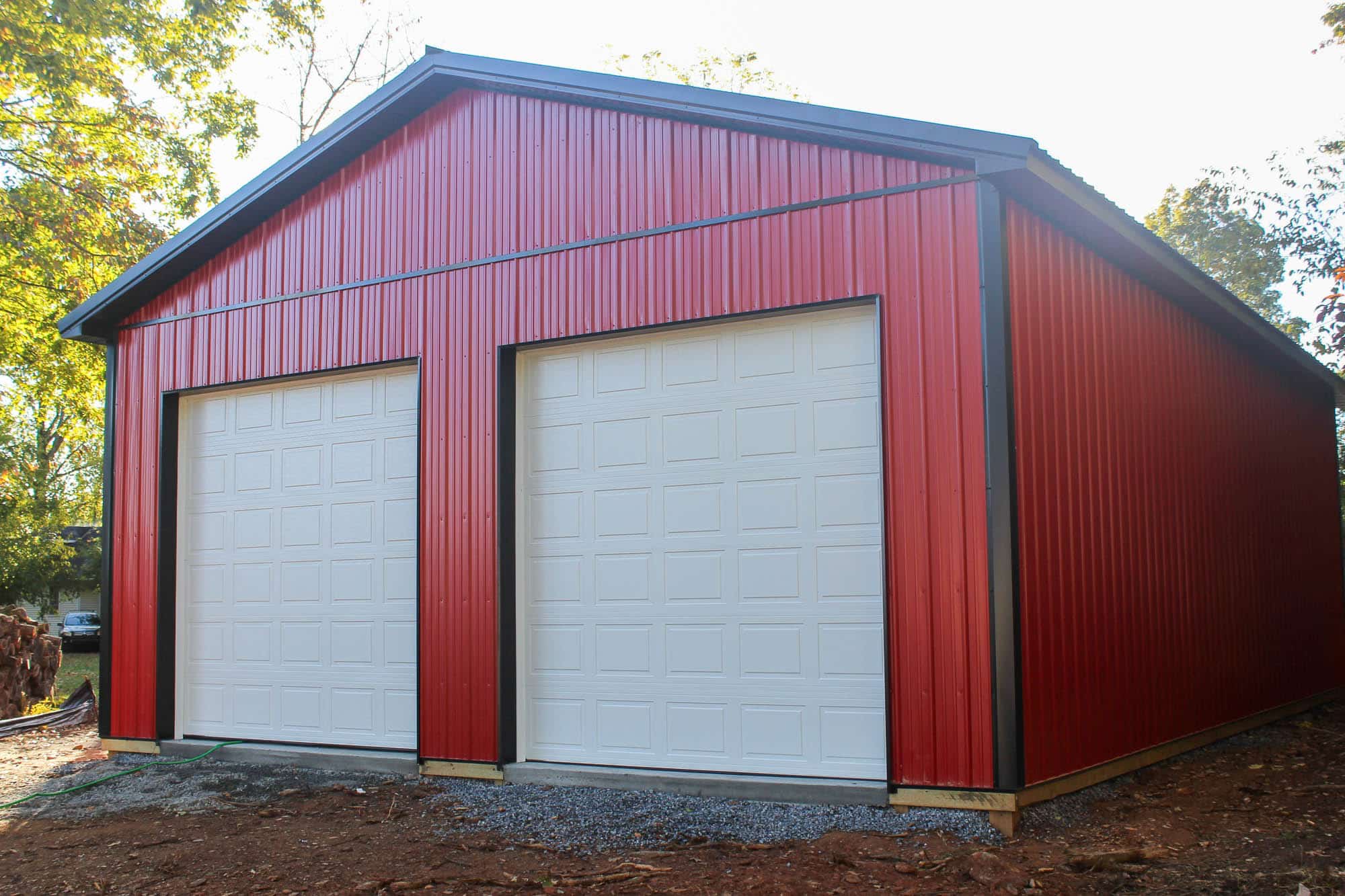 A red metal garage with a gable roof and two white roll-up garage doors. The structure is set on a gravel lot with a wooded area in the background.