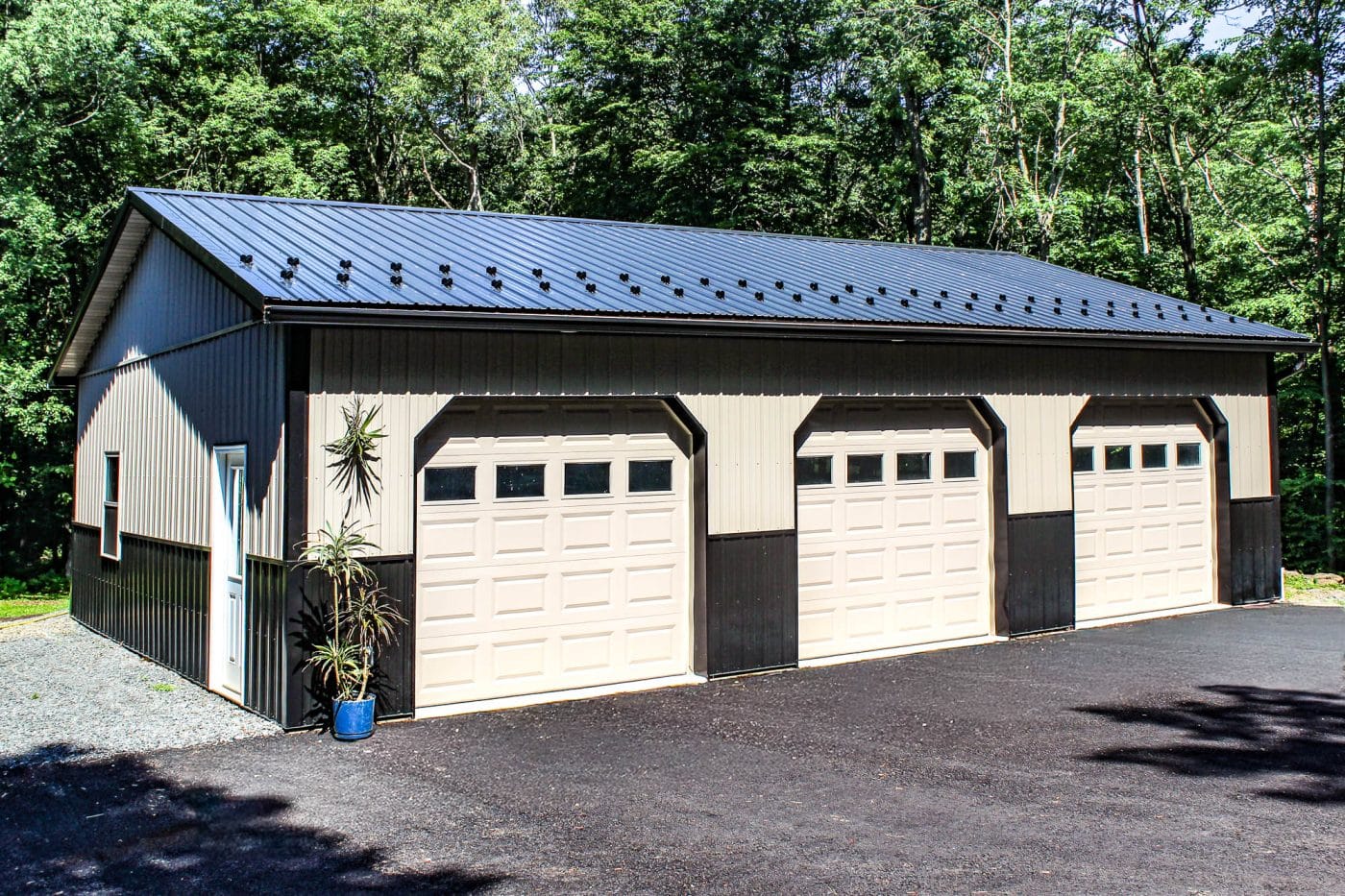 black and white three-bay post-frame garage with metal roof, overhead doors, asphalt driveway, and wooded background.