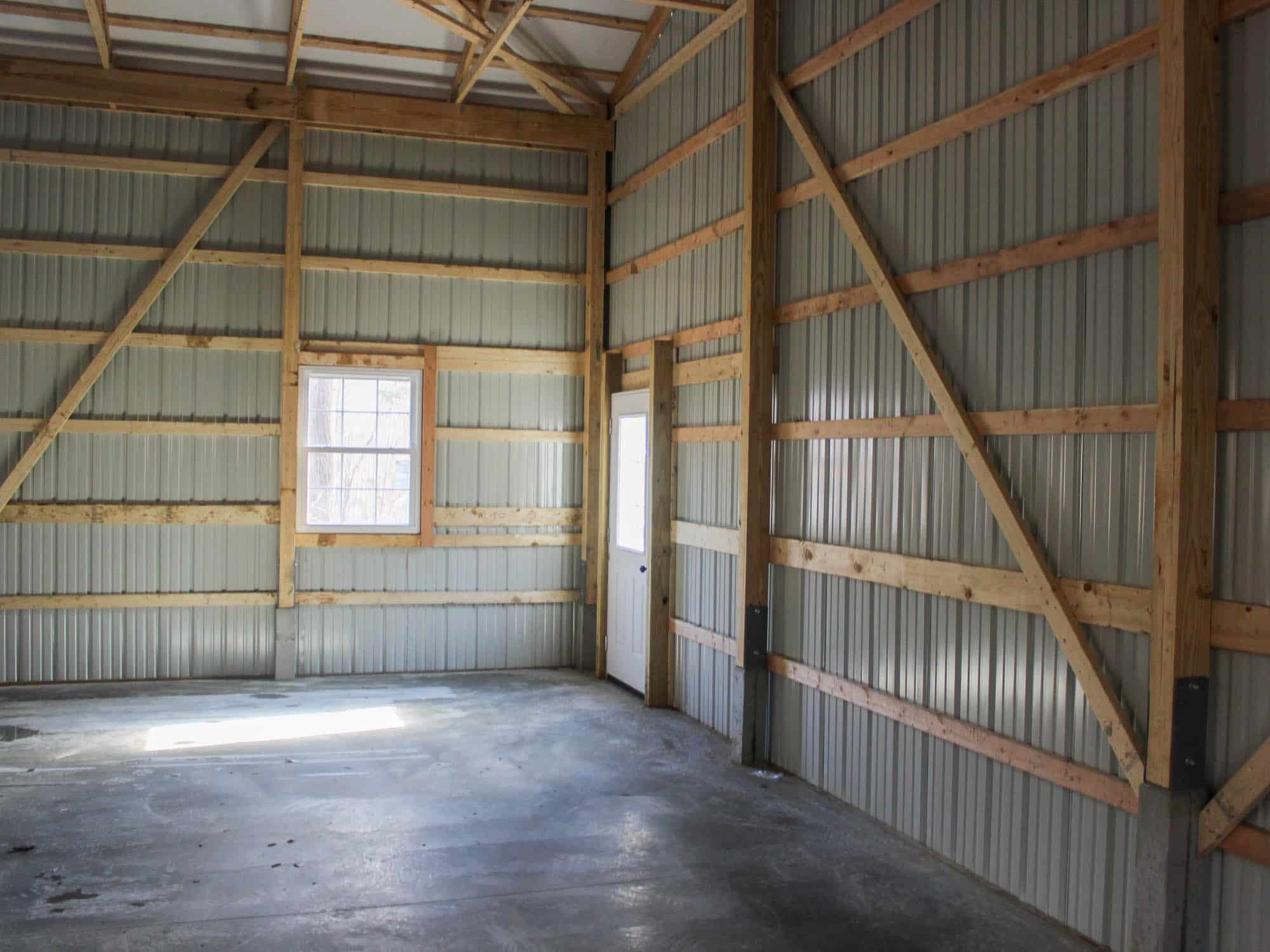 The interior of a pole barn featuring exposed wooden framing, metal siding, and a concrete floor. A single window allows natural light, and the roof is supported by wooden trusses.