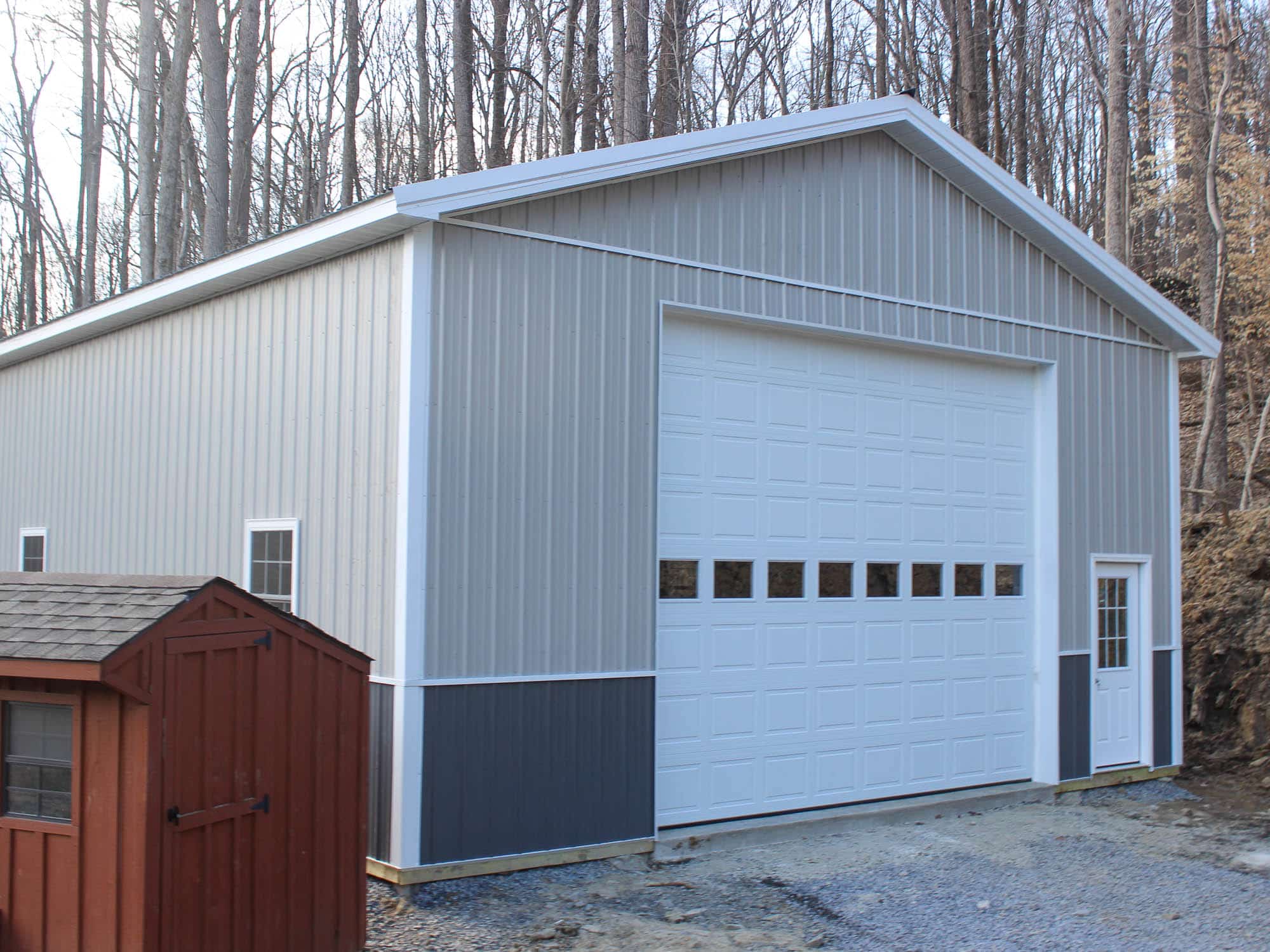 A gray metal pole barn with a large white roll-up garage door and dark gray wainscoting. The barn has a gable roof with white trim and is situated on a level gravel driveway with a wooded area in the background.