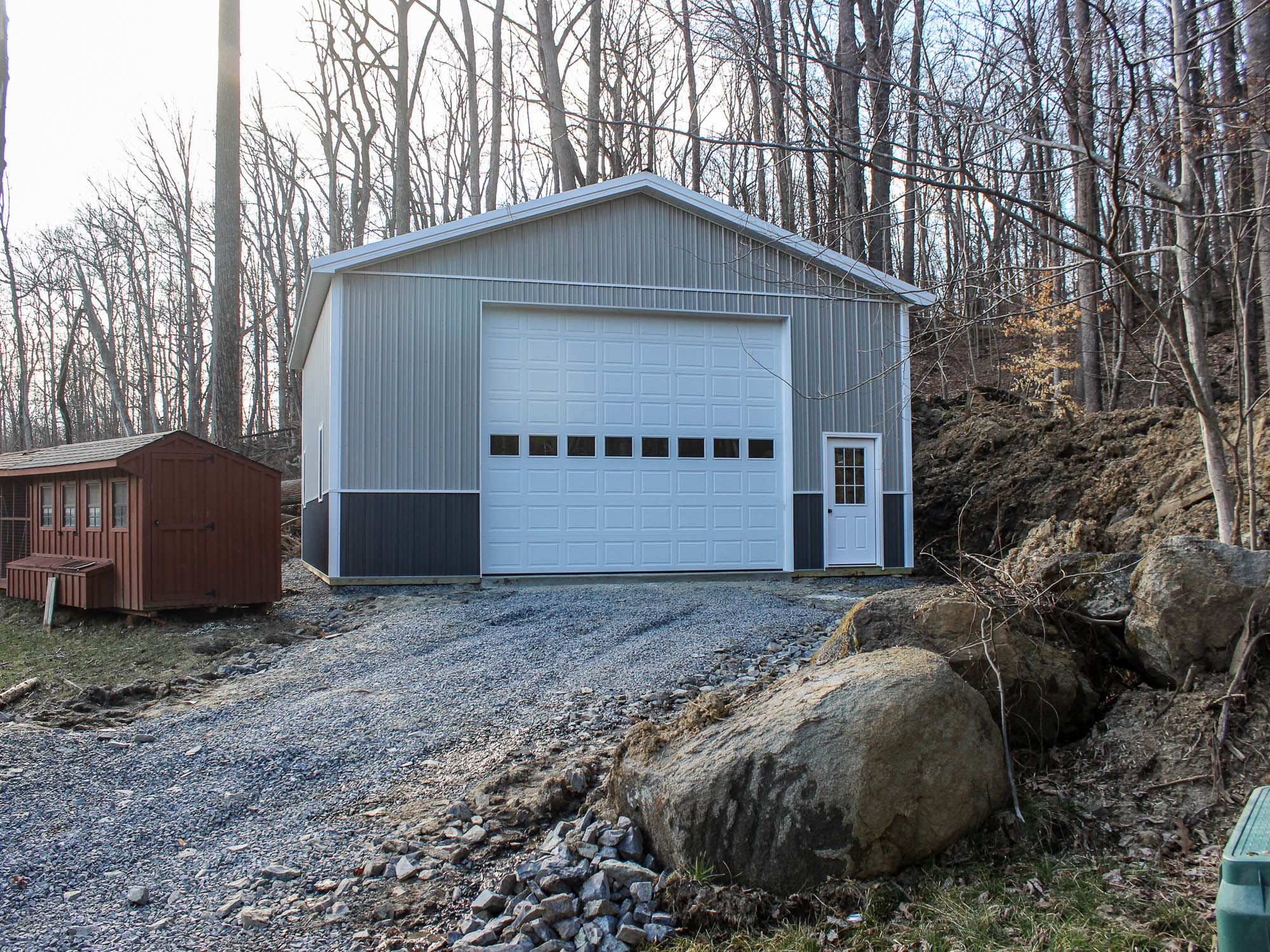 A gray metal pole barn positioned on an elevated gravel lot with a large roll-up garage door and a white side door. The building features dark gray wainscoting, a gable roof, and a small wooden shed nearby. A wooded hillside is visible in the background.
