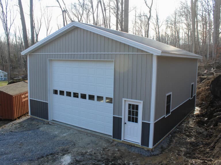 a gray metal pole barn with a large white roll-up garage door and a white side entry door. the barn has dark gray wainscoting along the lower section of the siding and a gable roof with white trim. a gravel driveway leads to the entrance, and a wooded hillside is in the background.