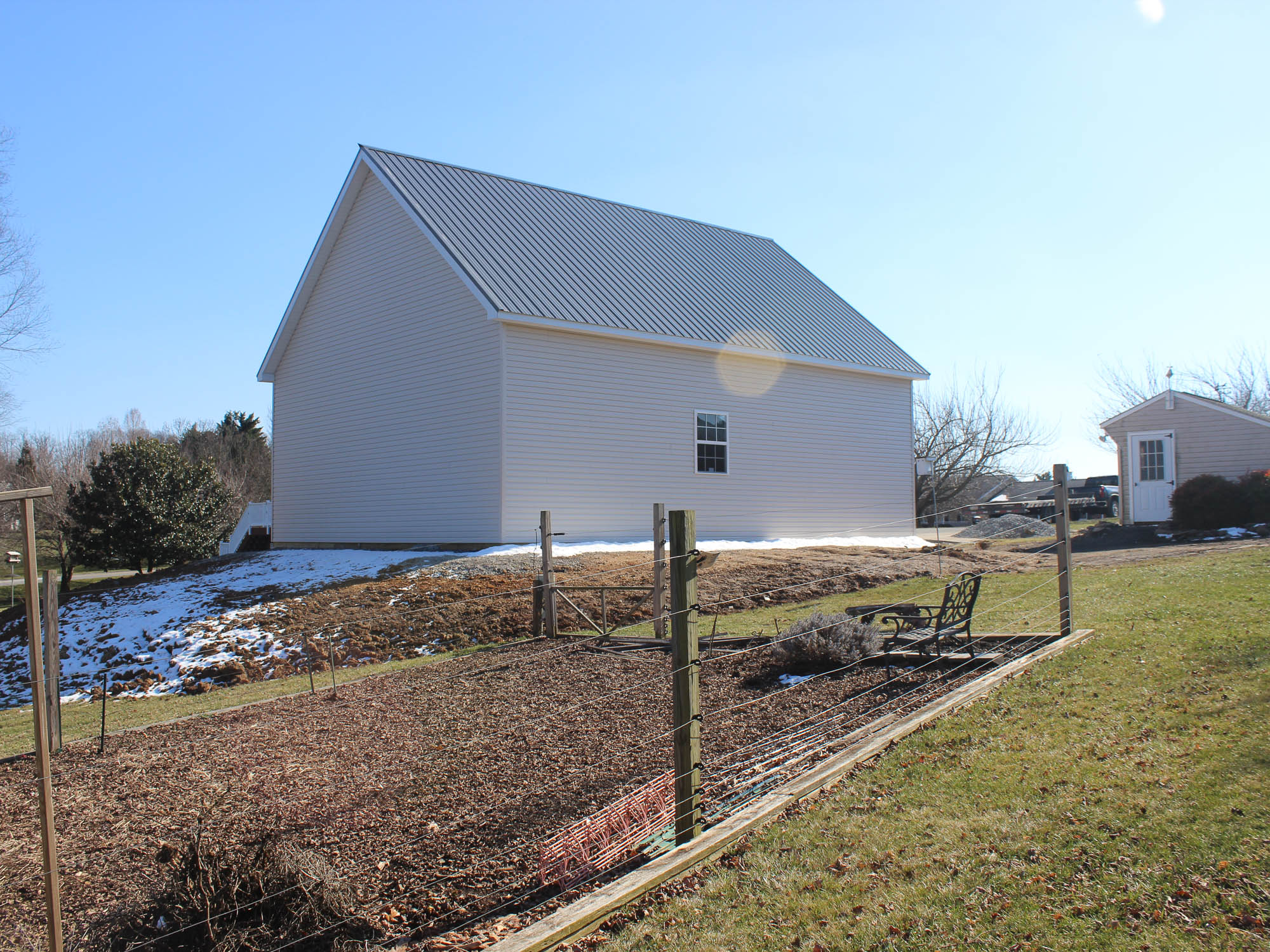 A white two-story garage with a gray metal roof and an open side yard. The structure is surrounded by grass and a gravel path.