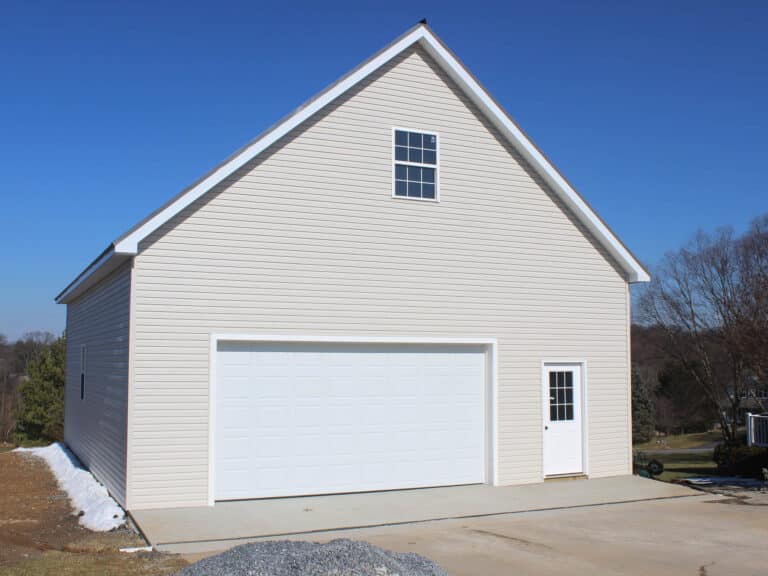 a front view of a white garage with a large white roll-up door and a gable roof. the garage sits on a concrete foundation with a gravel pathway leading to the entrance.