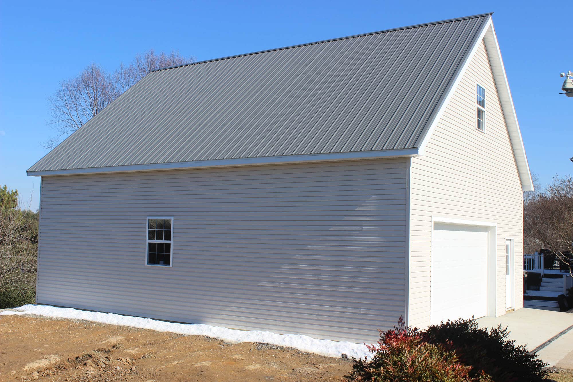 A side view of a white two-story garage with a gray metal gable roof. The building is located on a dirt lot with trees and neighboring houses in the background.