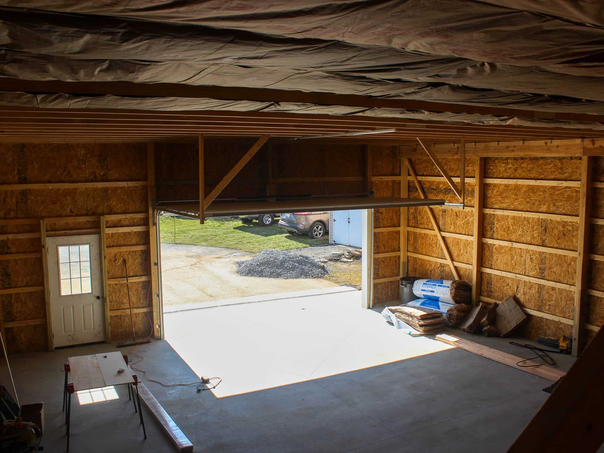 A lower-level interior view of a garage with a workbench, a concrete floor, and an open garage door. Natural light streams in from the door.