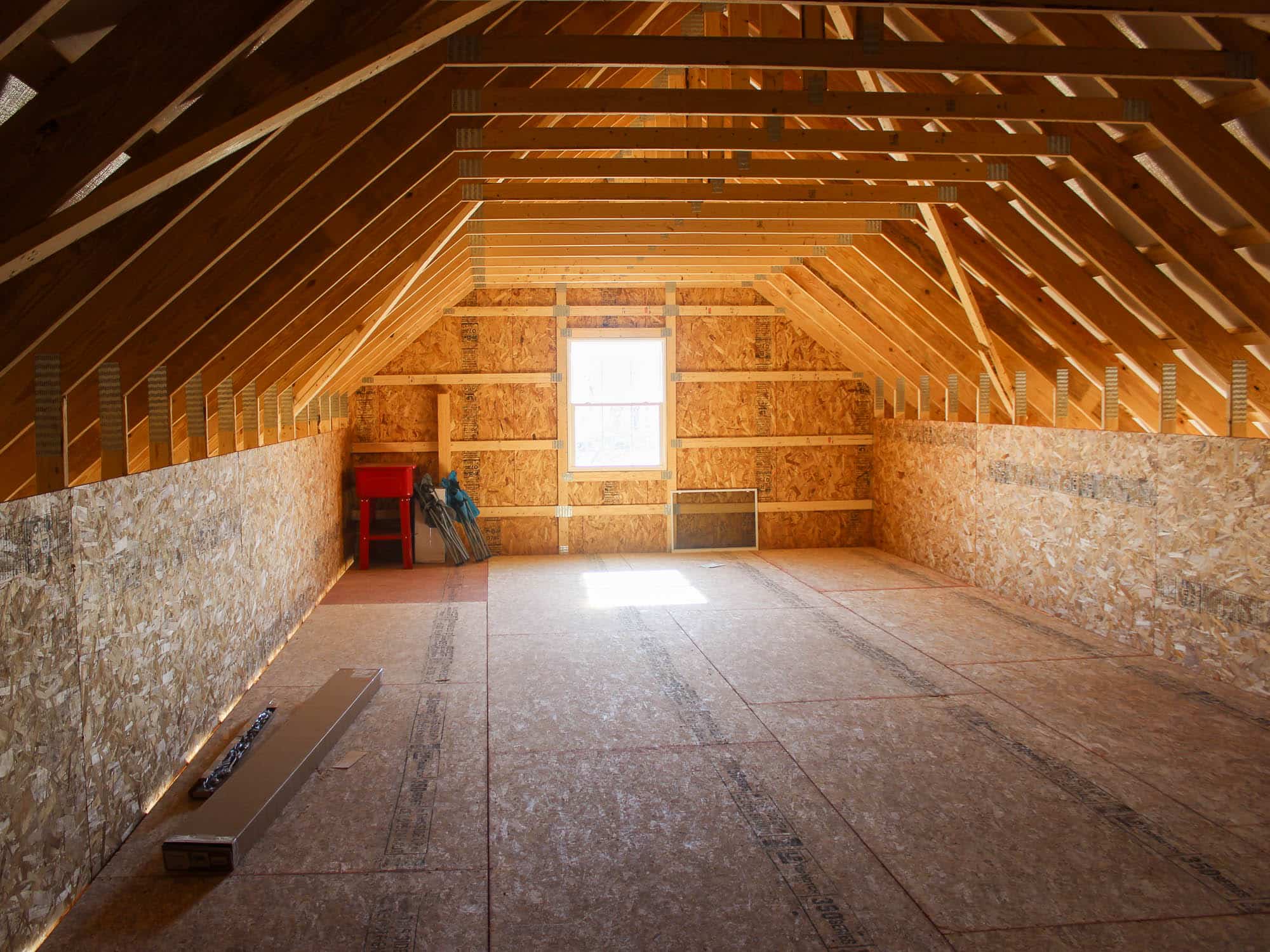 The interior of a two-story garage featuring an open loft with exposed wooden rafters and plywood flooring. A single window allows natural light.