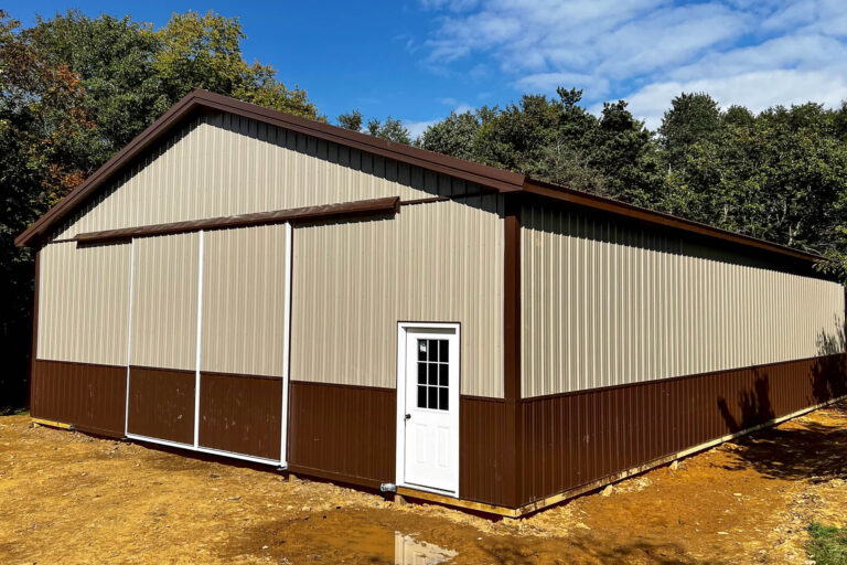 a beige metal building with dark brown wainscoting, a white side entry door, and a gable roof with dark trim. the structure is positioned on a gravel lot with trees in the background.