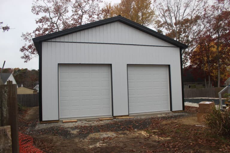 a gray metal garage with dark gray trim, a gable roof, and two white roll-up garage doors. the building sits on a gravel driveway with trees and grass in the background.