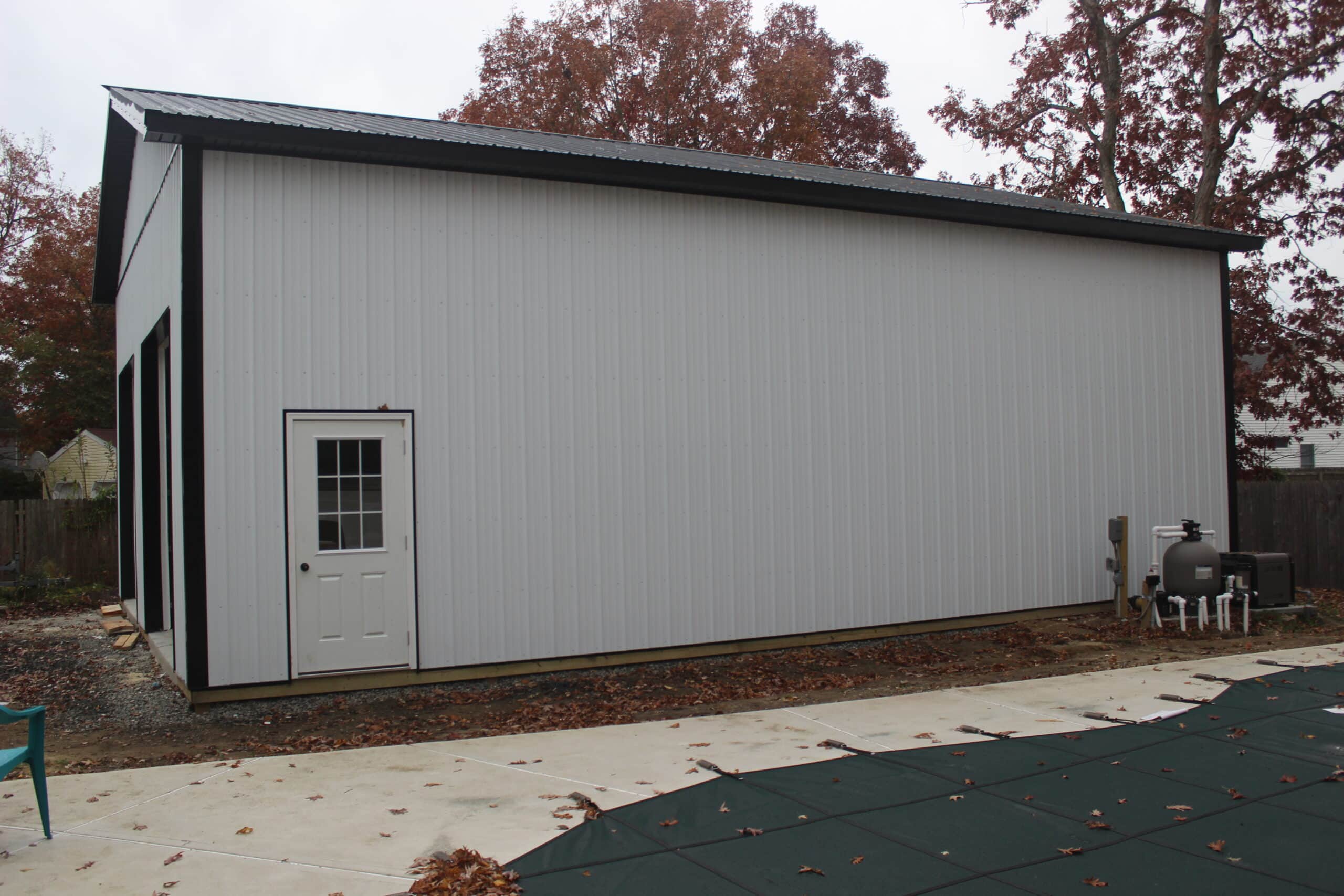 A side view of a gray metal garage with a gable roof, dark trim, and a single white entry door. The structure sits on a paved driveway with trees in the background.