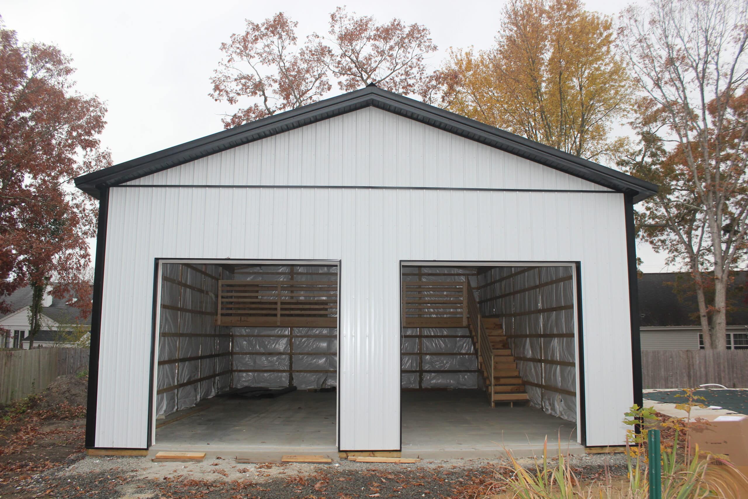 A side view of a gray metal garage with a gable roof, dark trim, and a single white entry door. The structure sits on a paved driveway with trees in the background.