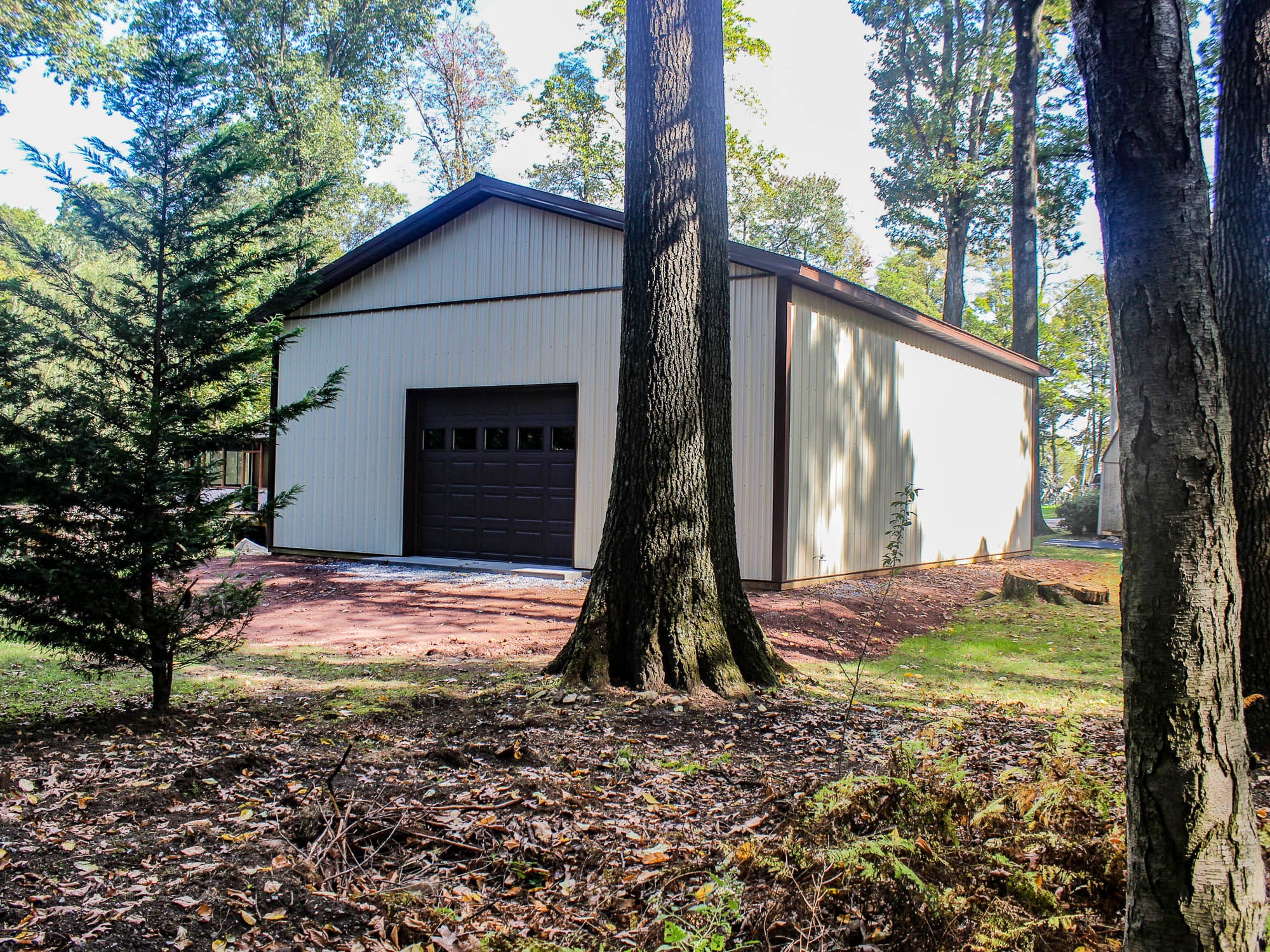 A beige metal garage with dark brown trim, featuring a large roll-up door and a gable roof. The structure is nestled among tall trees, with a forested area in the background and a gravel path leading to the entrance.