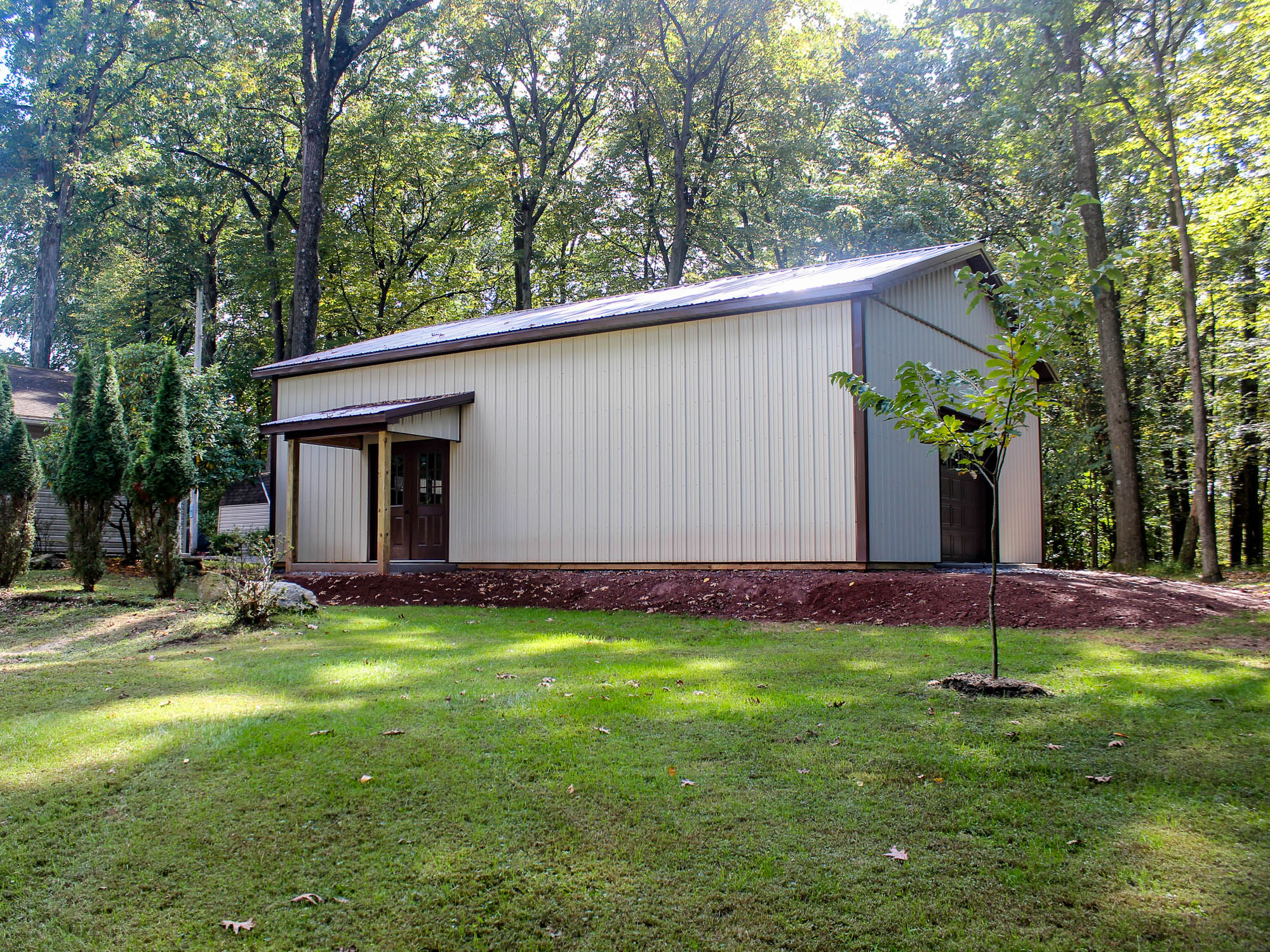 A beige metal workshop with a brown trim gable roof, featuring a side entry door and a large garage-style door. The building is surrounded by a well-maintained grassy lawn with tall trees in the background.