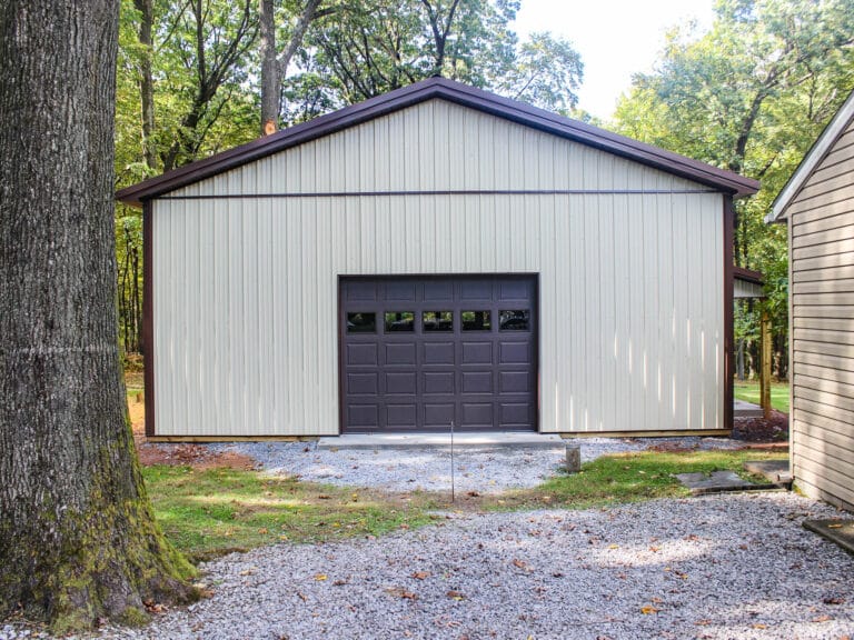 a beige metal garage with a dark brown roll-up door, situated in a wooded area with a gravel driveway leading up to the entrance. the building features a simple gable roof with dark brown trim, blending with the surrounding trees.