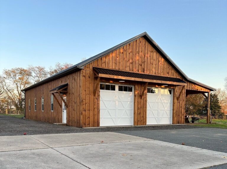 a 32'x60' wooden barn with two white garage doors and a covered entryway. the barn features vertical wood siding, a dark metal roof, and exposed wooden beams under a clear sky.