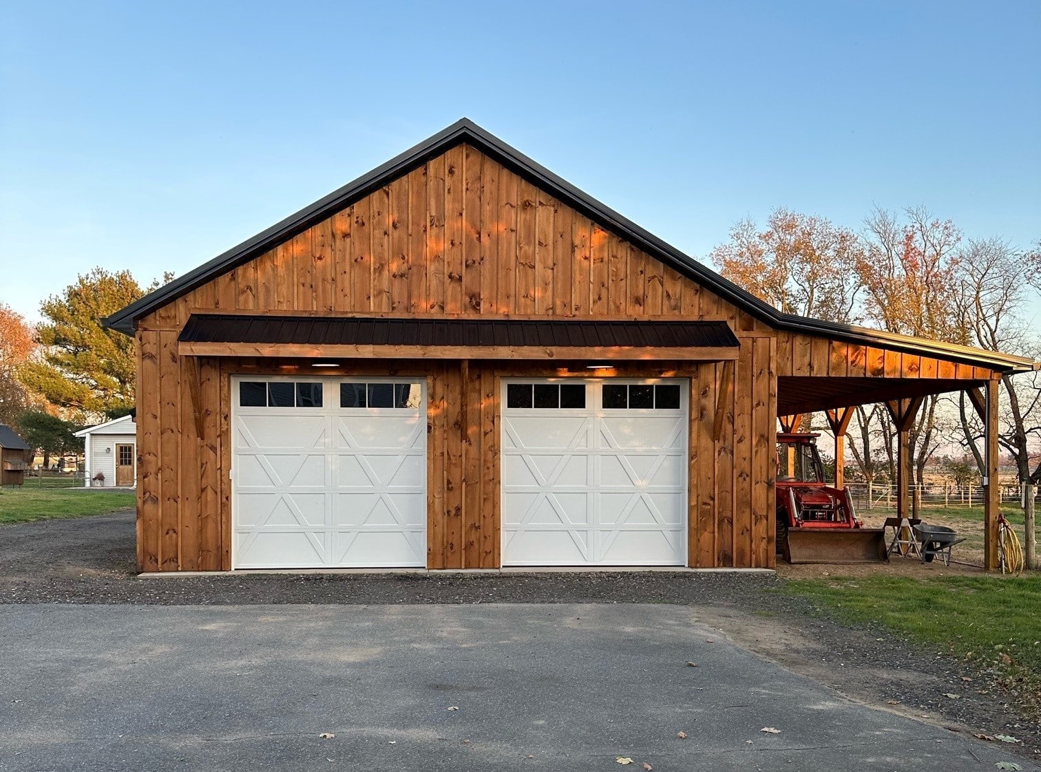 A front view of a wooden barn with two white garage doors and a side entry with a covered porch supported by wooden posts.