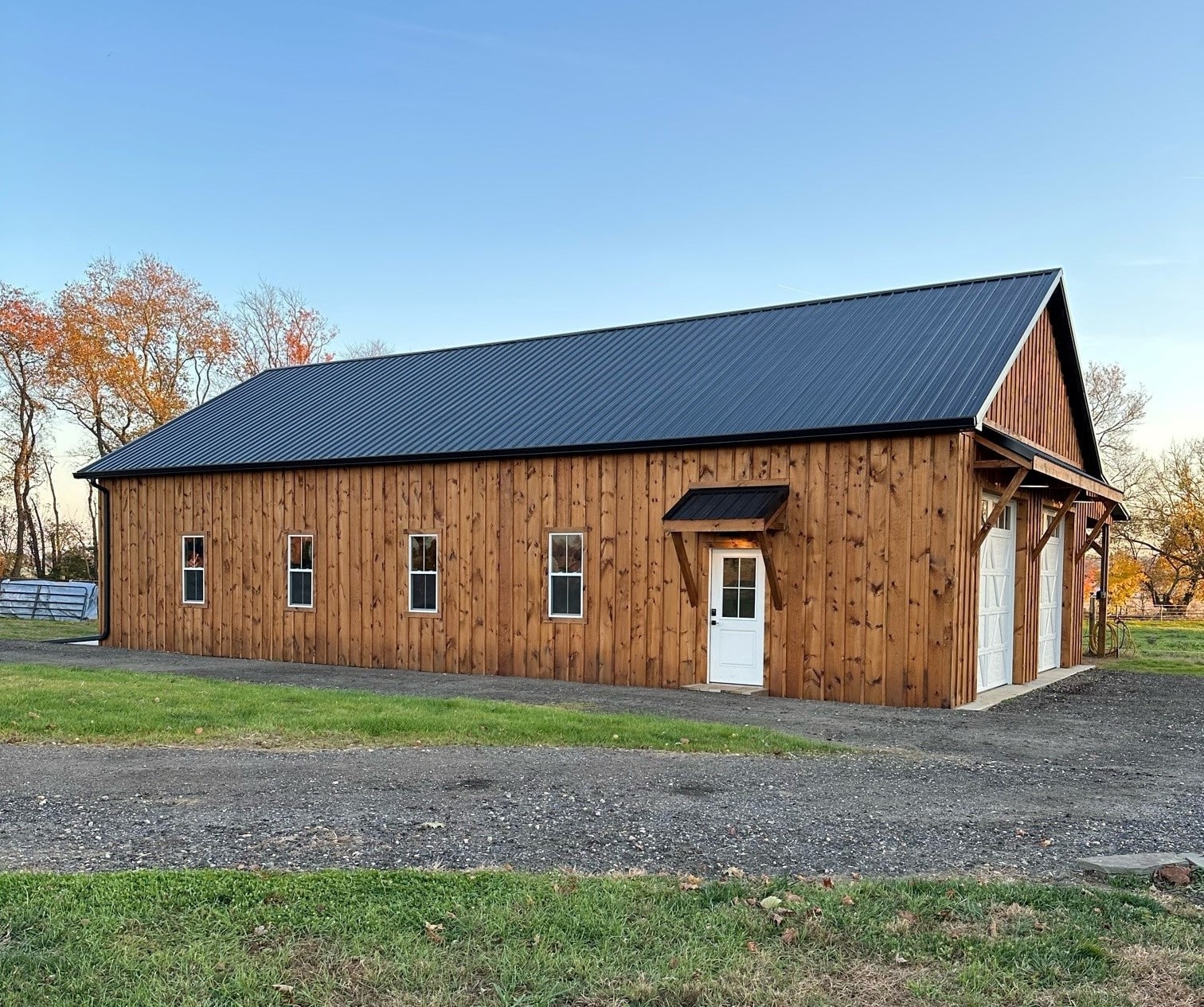 A wooden barn with a side entry door, several windows, and a black metal roof. The building is set on a gravel base with a grassy field in the background.