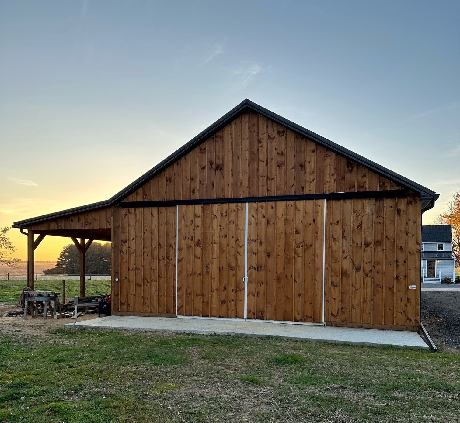 A back view of a wooden barn with large sliding wooden doors and a covered side area. The barn features a black metal roof and vertical siding.
