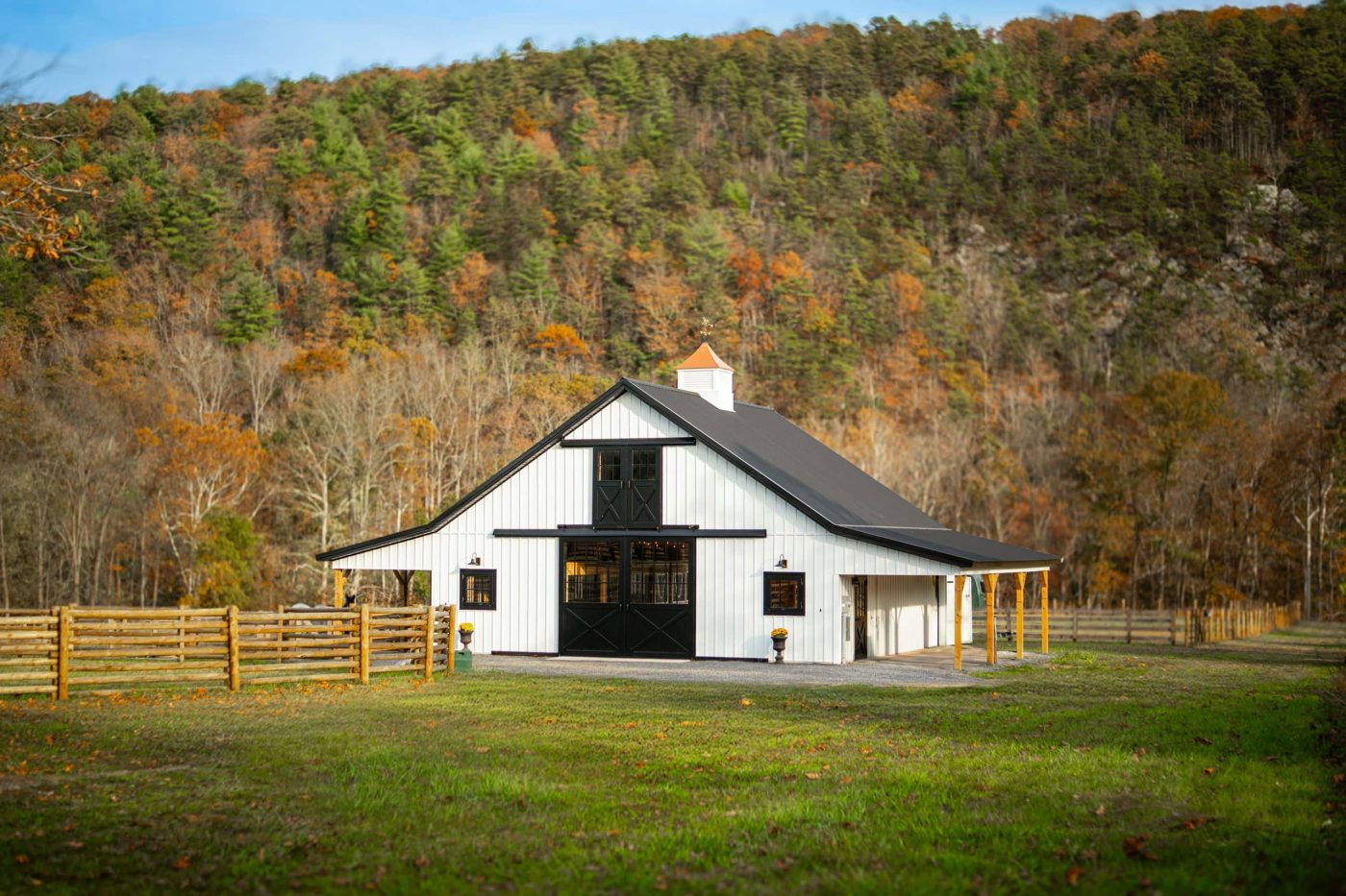 white horse barn with black trim, a cupola, and an attic loft, featuring board and batten siding and a metal roof in a rural autumn landscape.