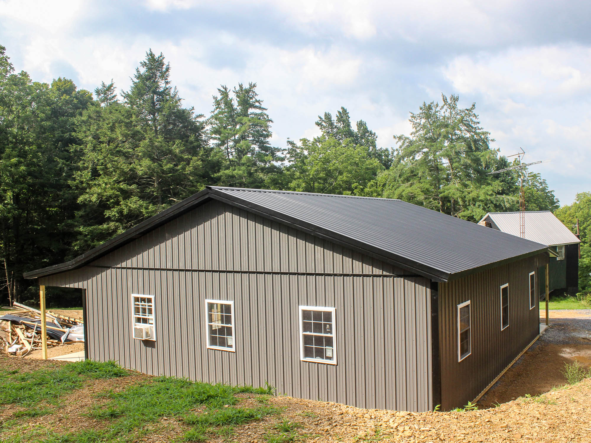 A side view of a gray metal building with a black gable roof and multiple white-trimmed windows. The building sits on a gravel foundation.