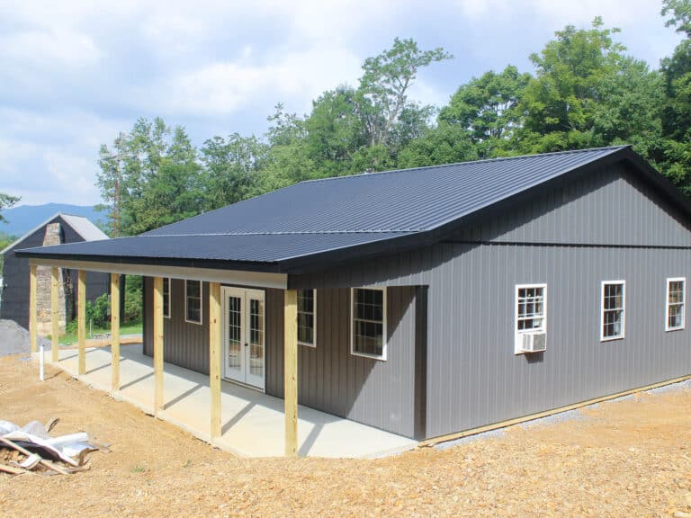 an angled view of a gray metal building with a black gable roof, a covered porch supported by wooden posts, and side windows.