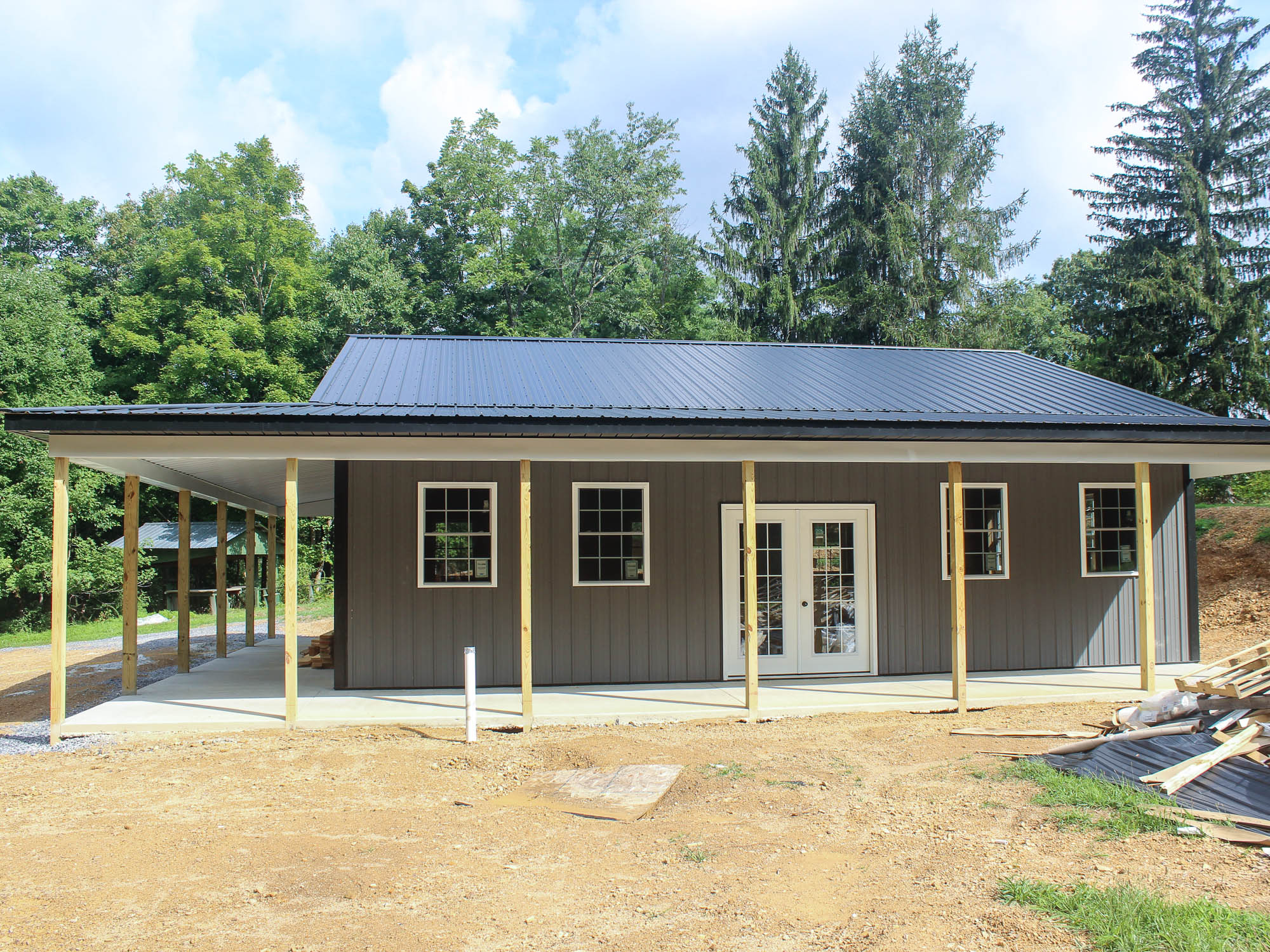 A front view of a gray metal building with a gable roof, two glass doors, and several windows. The building sits on a dirt lot with trees in the background.