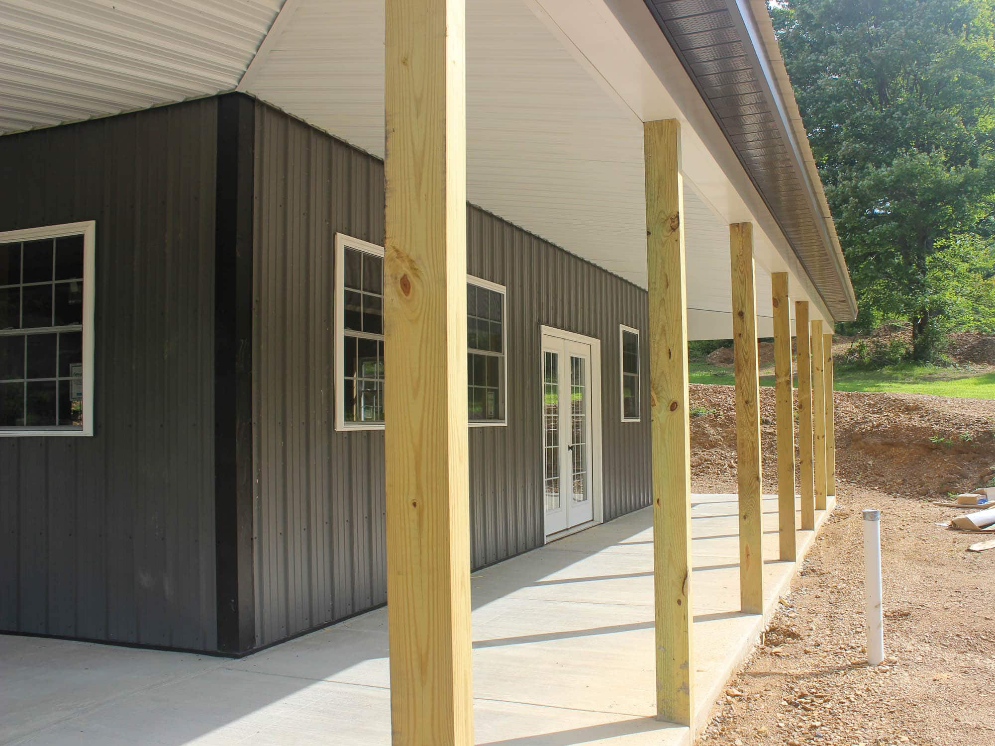 A close-up of the covered porch of a gray metal building, showing wooden posts and concrete flooring. A single window and door are visible under the porch roof.